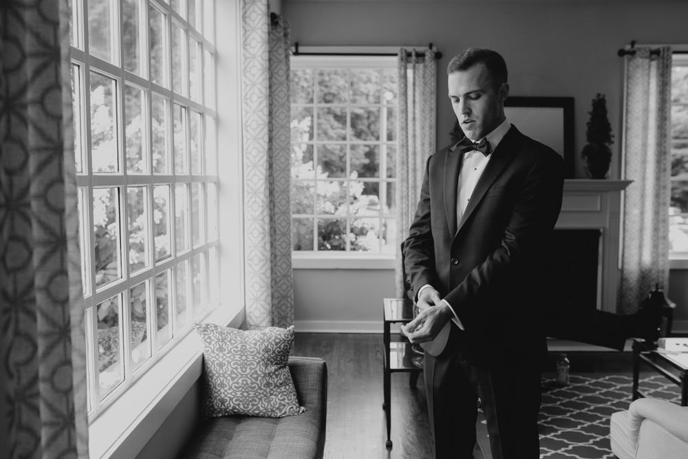 A black and white photo of someone in a tuxedo standing by a large window in an elegant room.
