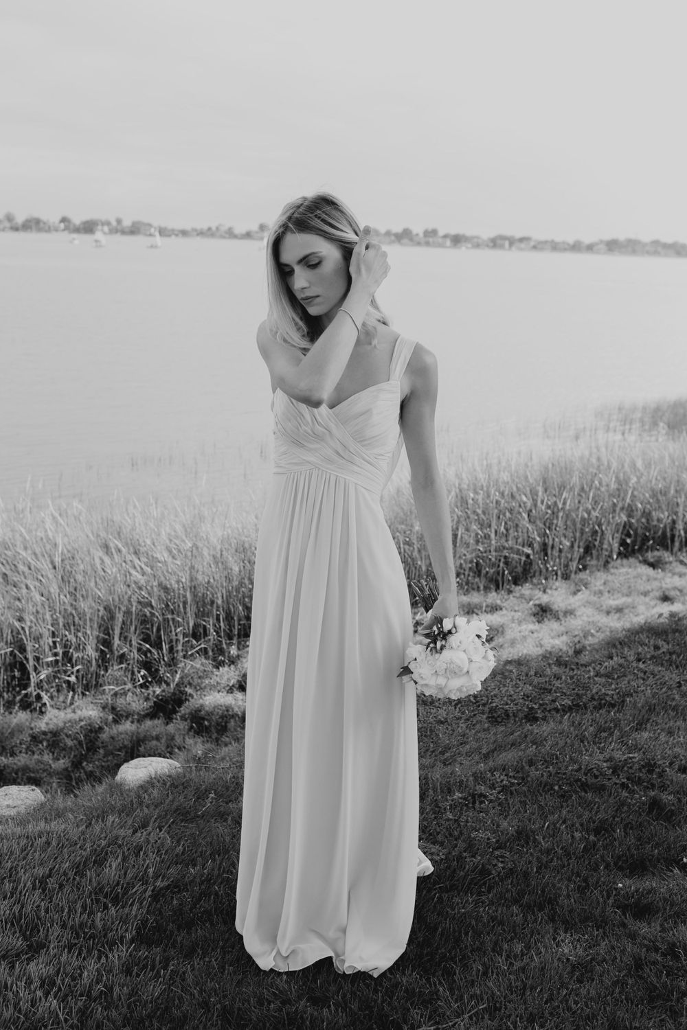 Black and white portrait of bride in elegant dress holding bouquet by waterfront.