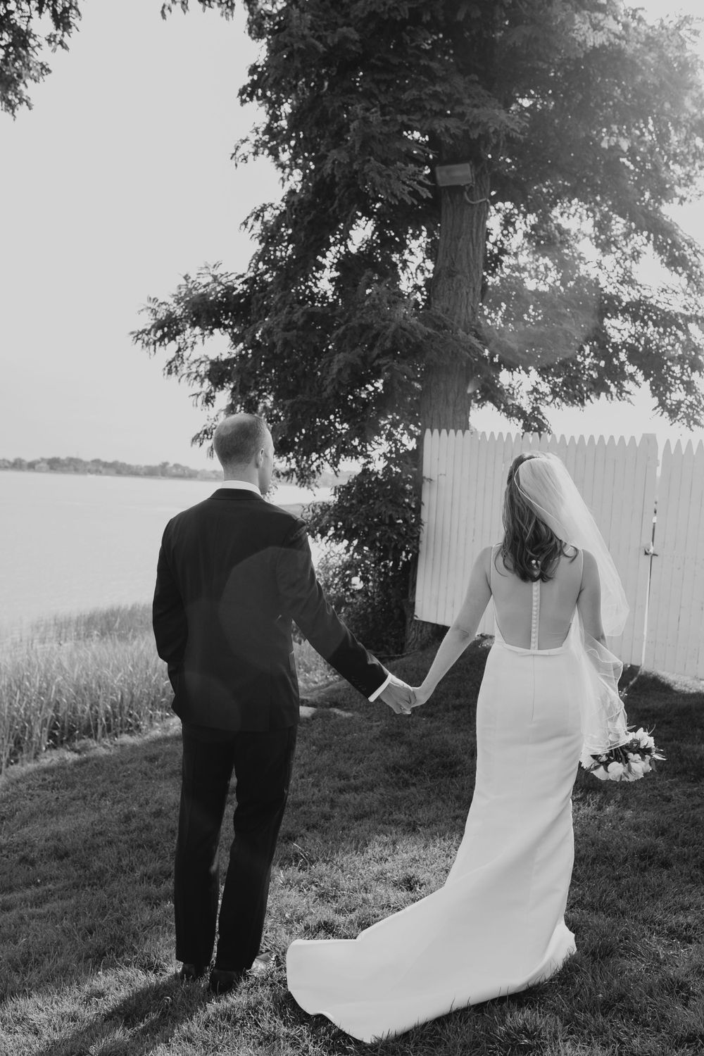 Newlyweds hold hands while walking together by water's edge in black and white photo.