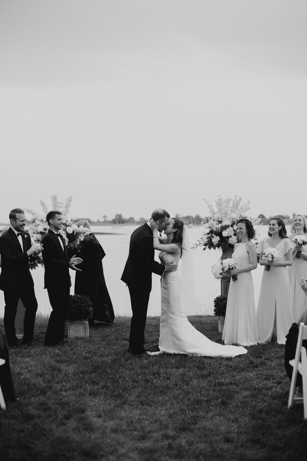 Black and white wedding ceremony photo with wedding party lined up on grassy waterfront.