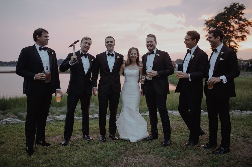 Wedding party posing with drinks against sunset waterfront backdrop.