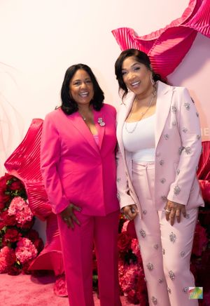 Two women pose together wearing pink and light pink suits against floral backdrop.
