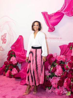 Woman in white blouse and pink pleated skirt stands against pink floral wall.