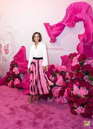 Woman in white blouse and pink pleated maxi skirt poses against pink floral backdrop.