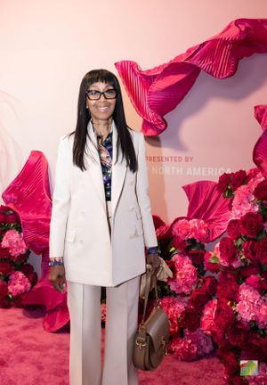 A guest in a white suit jacket and glasses poses against a pink backdrop with red floral decorations.