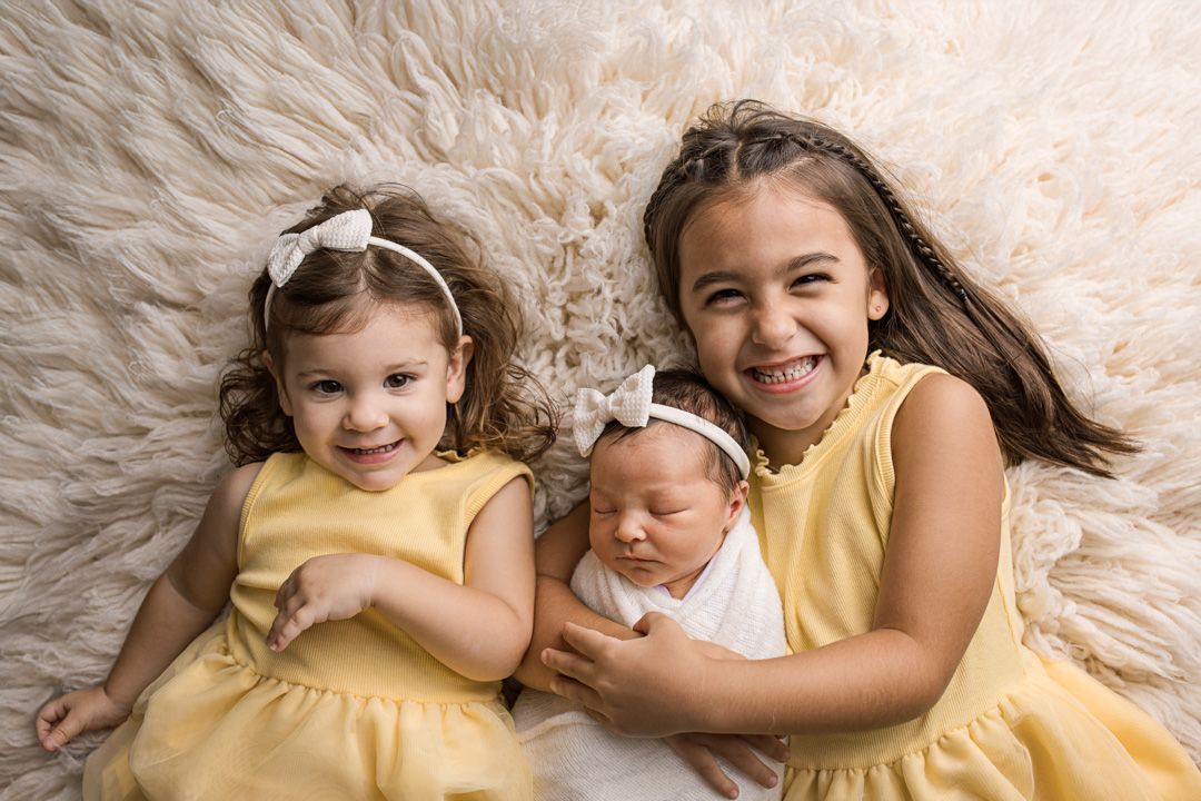 Three siblings wearing yellow dresses pose together on a cream-colored furry backdrop during a professional photo session.
