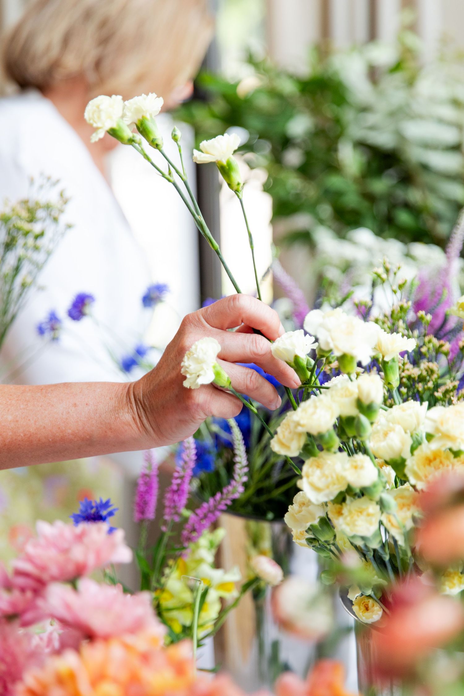 Hands arranging white carnations and colorful flowers in a bright, airy setting.