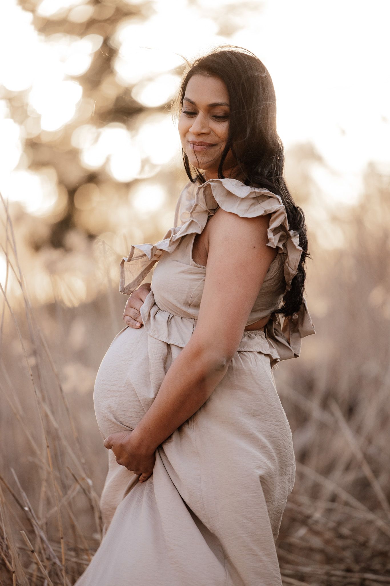 Pregnant woman in flowing dress smiles while cradling her belly in warm sunset light.