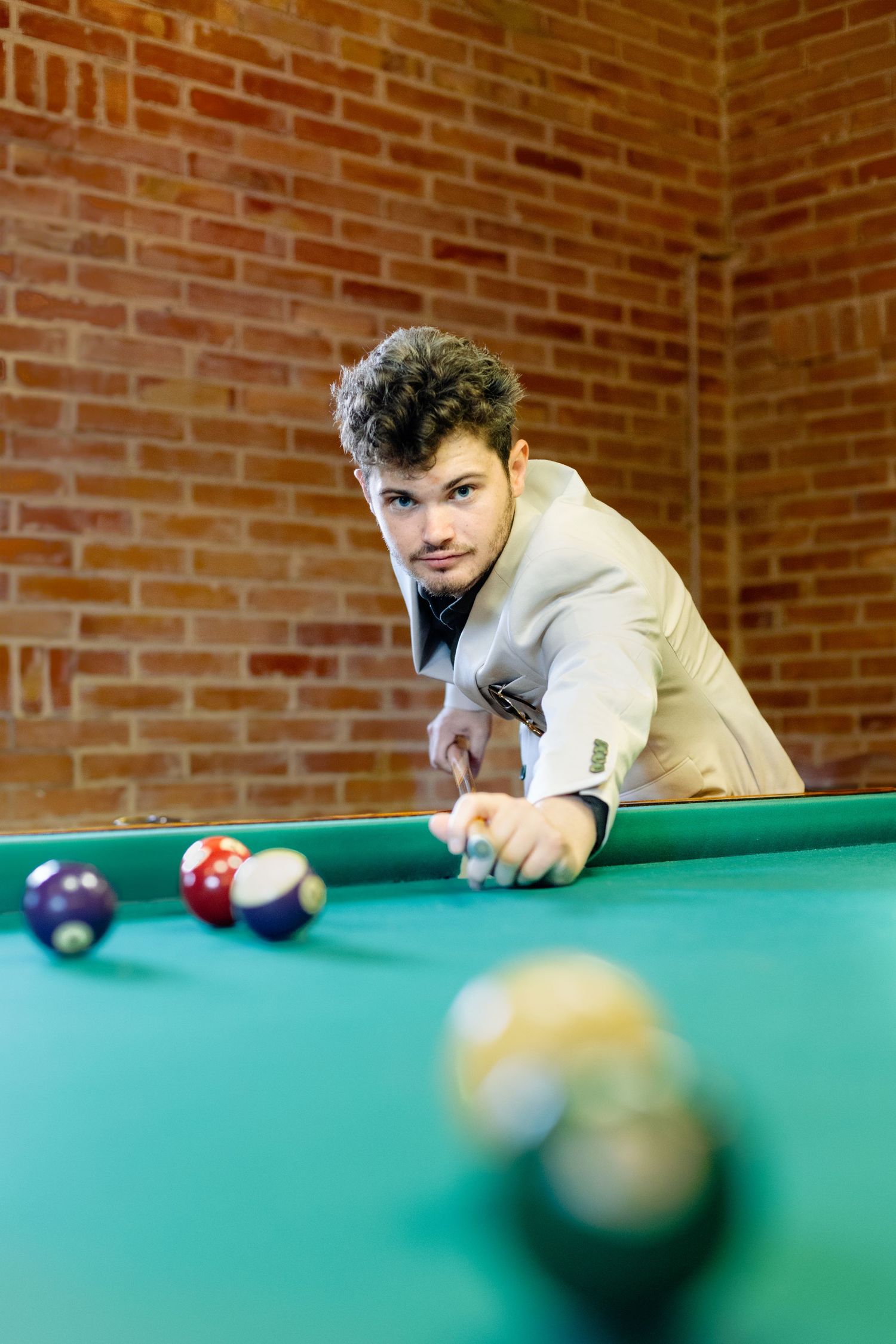 Person in beige suit jacket leans over green pool table preparing to take a shot at billiards balls.