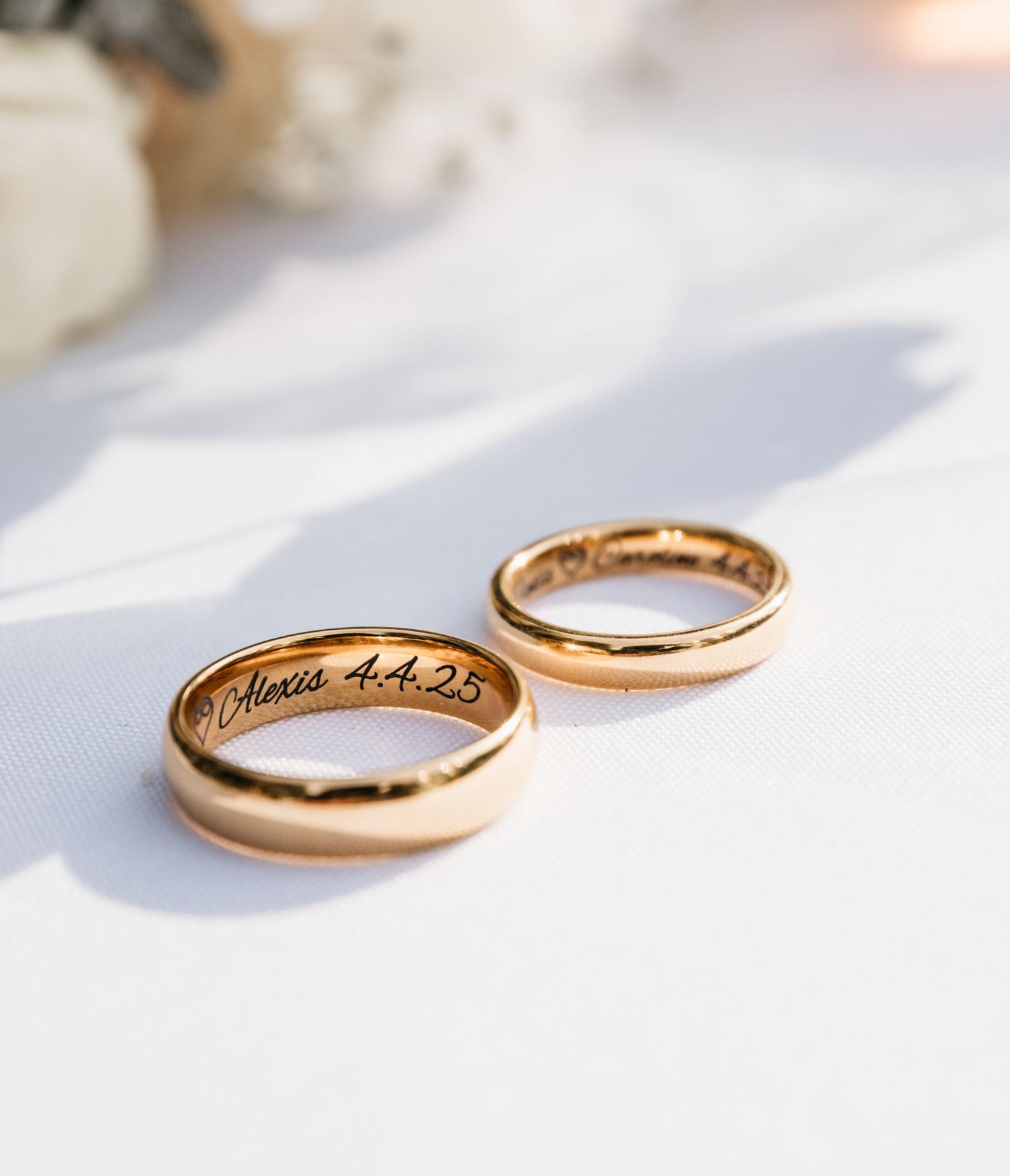 Two gold wedding rings with engraved inscriptions resting on a white surface casting soft shadows.