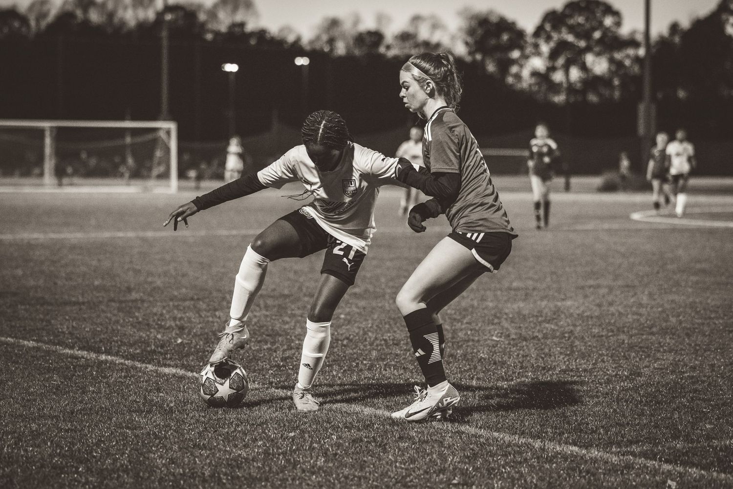 Black and white photo of two female soccer players competing for ball during night game.