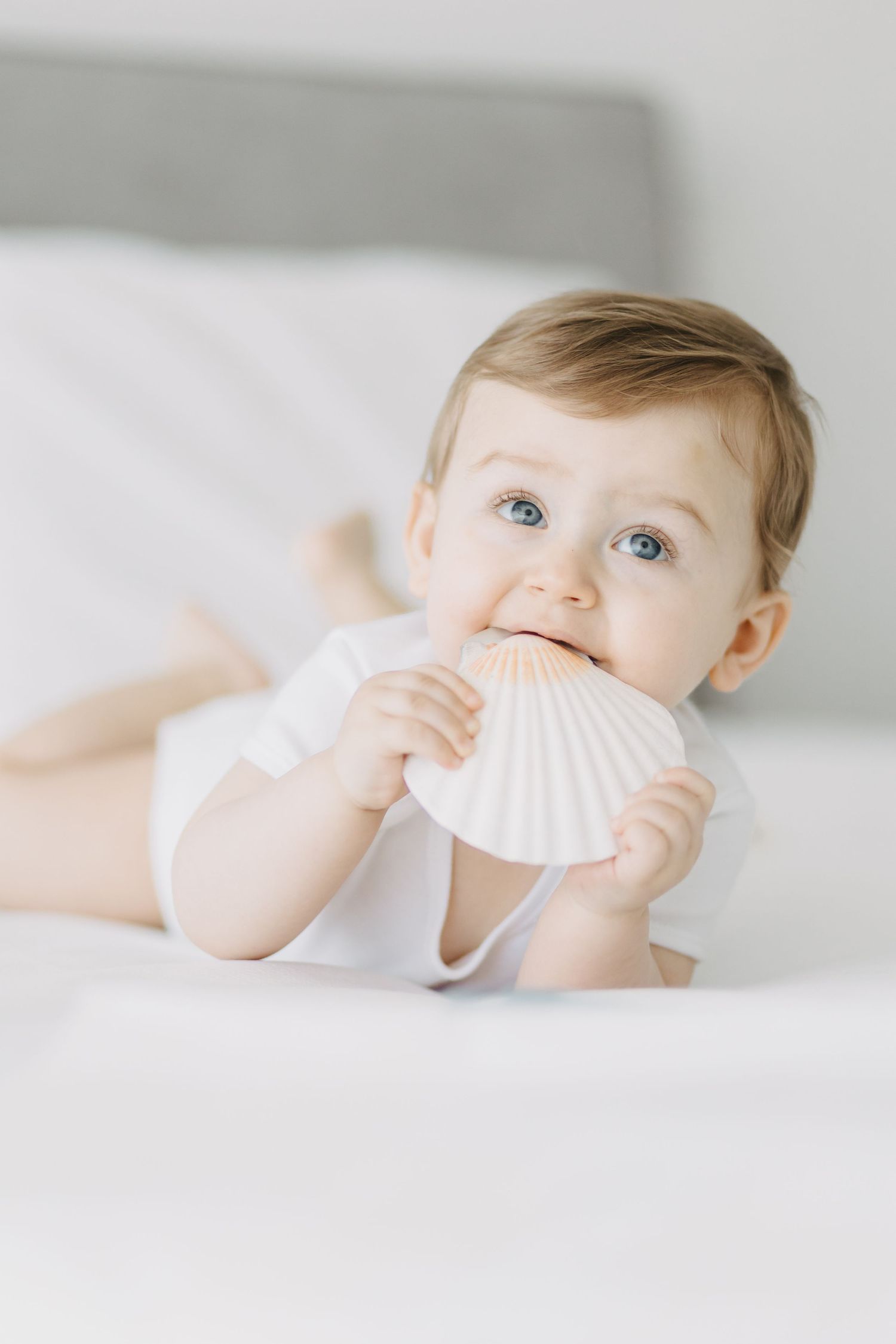 Baby lying on white bedding playing with seashell.