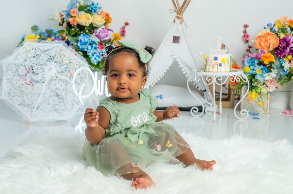 A baby girl in a green tulle dress sits on a fluffy white rug surrounded by colorful flowers and a teepee backdrop.
