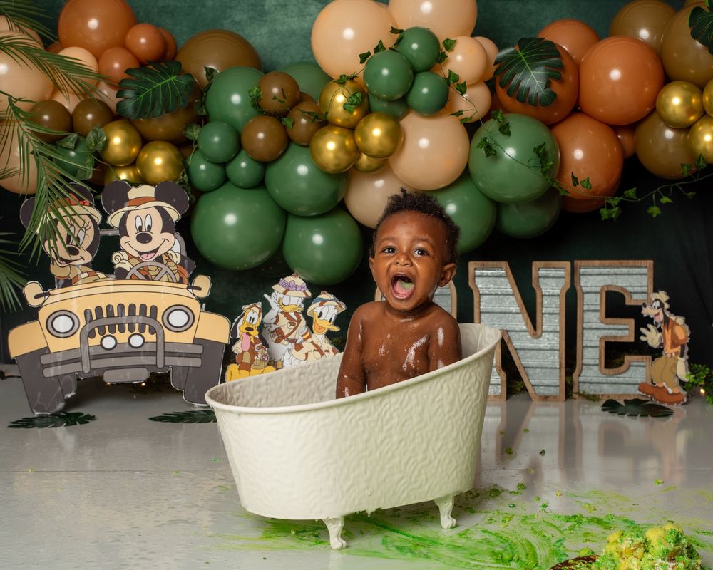 Baby's first birthday cake smash in a white tub with Mickey Mouse theme, green balloons, and ONE sign backdrop.