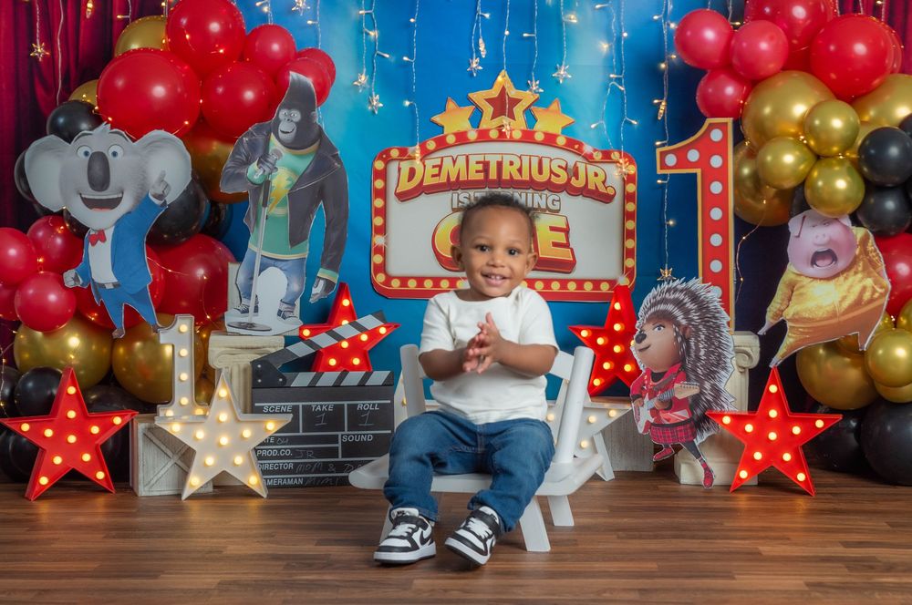 A toddler boy sits in a white chair at a Sing-themed first birthday party with colorful balloons and character cutouts.
