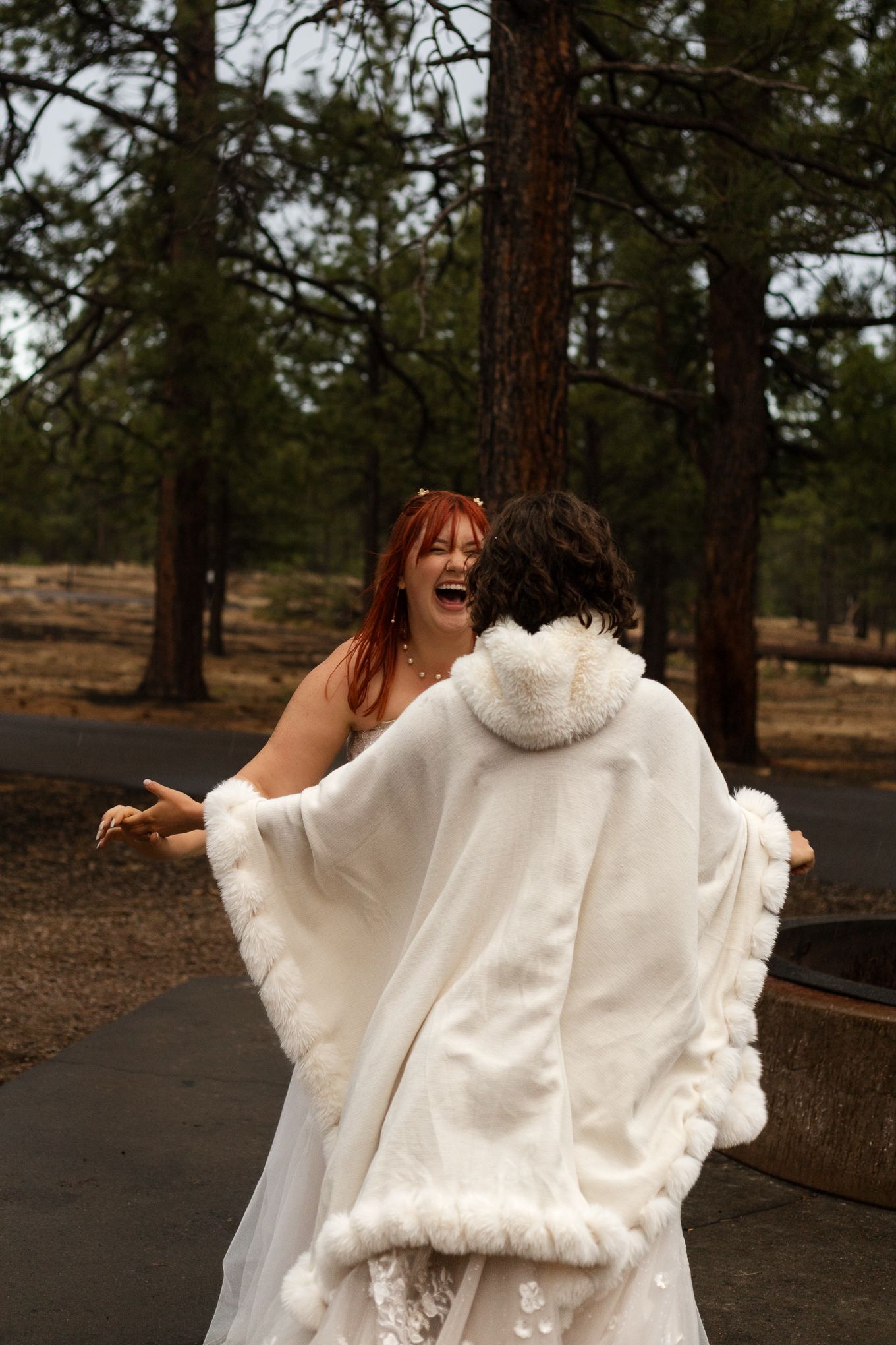 Two people in white wedding attire laugh joyfully together among pine trees in a forest setting.