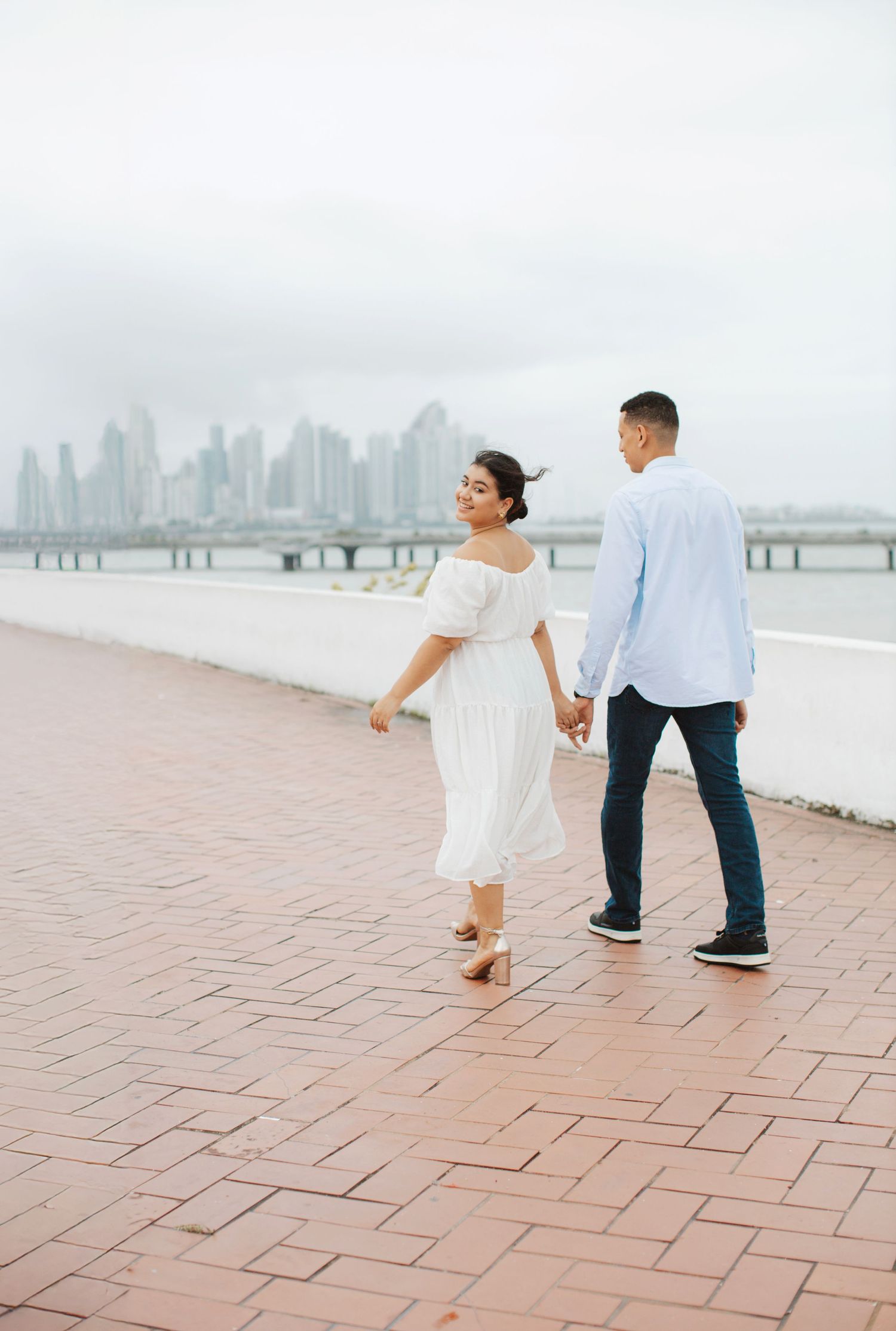 Couple walking hand in hand along waterfront with city skyline in background.