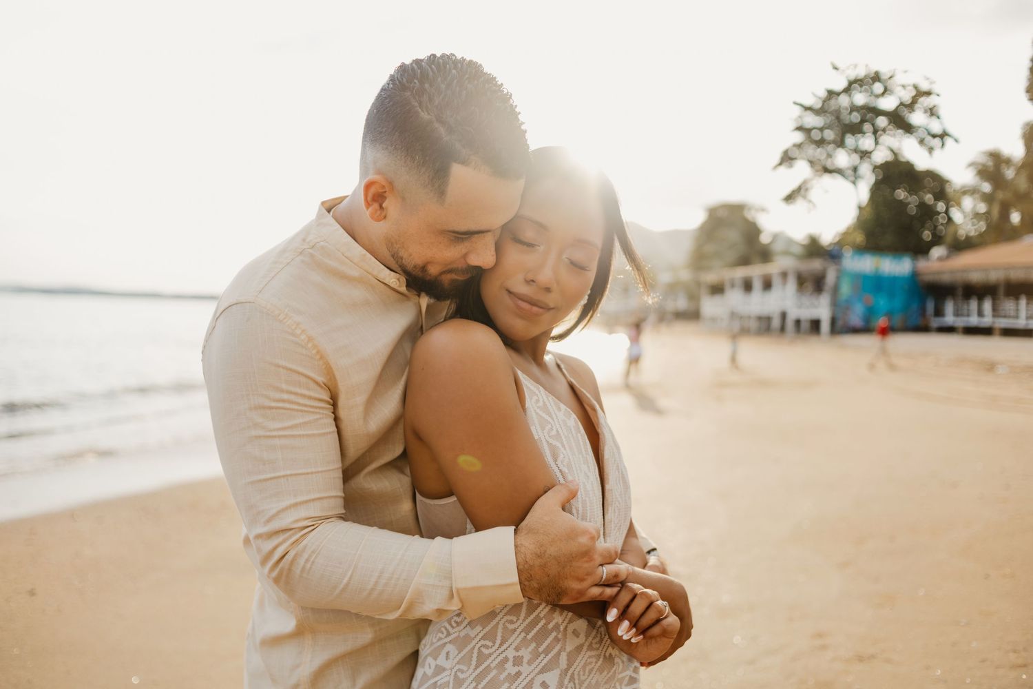 Romantic sunset beach embrace between couple in light colored outfits on sandy shore.