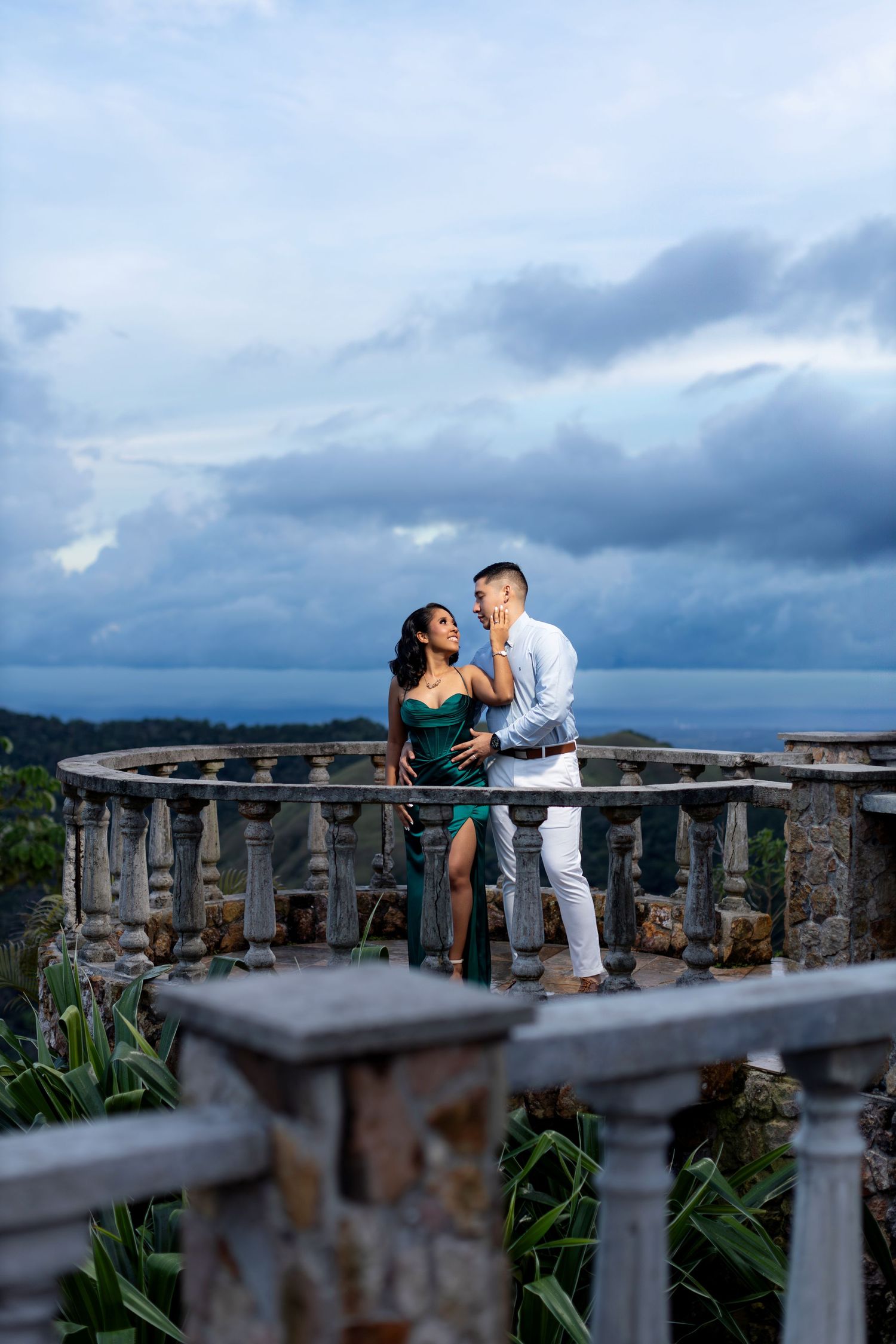 Romantic couple poses on stone balcony overlooking mountain vista at sunset.