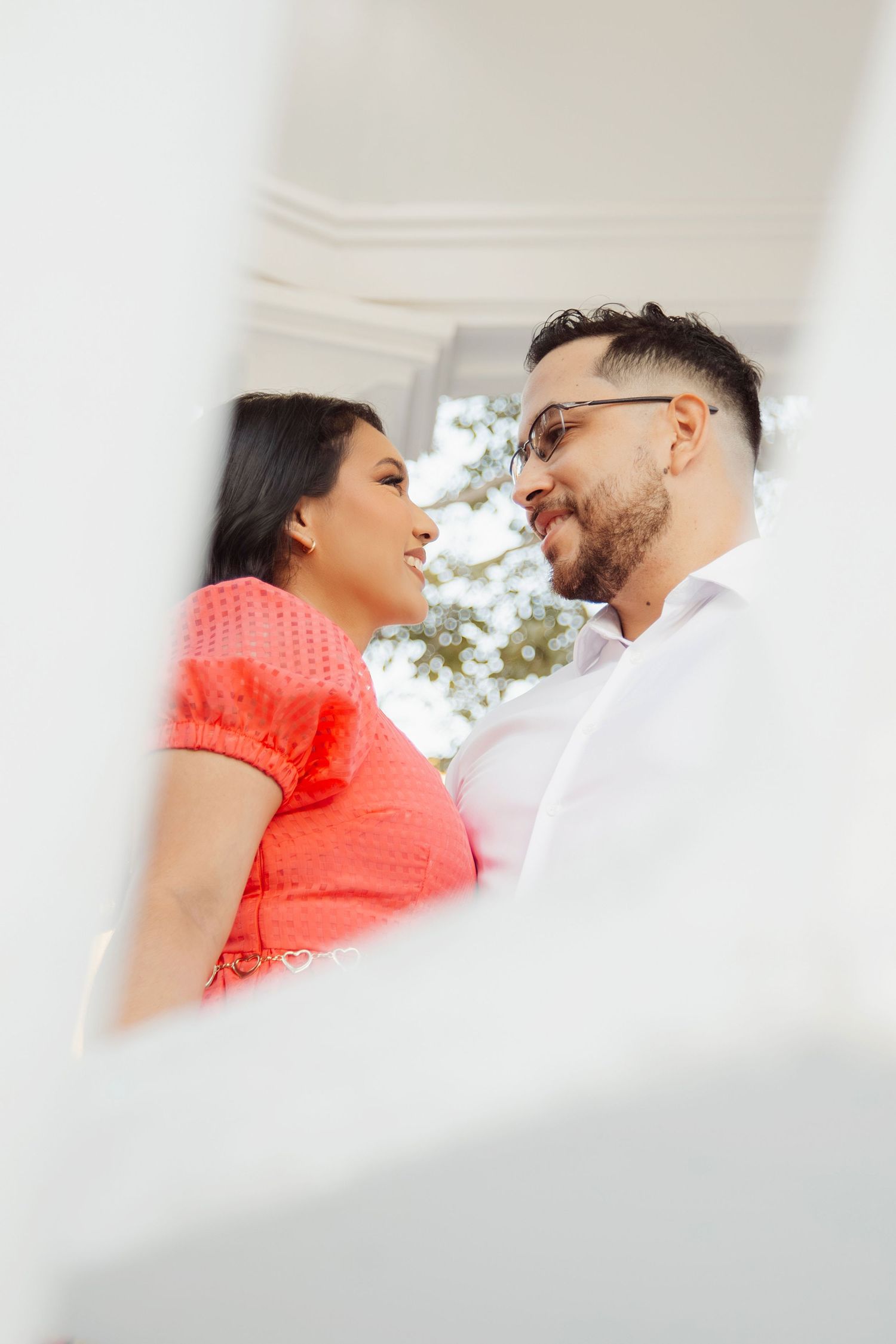 Romantic couple in white and coral outfits sharing intimate moment framed through architectural detail.