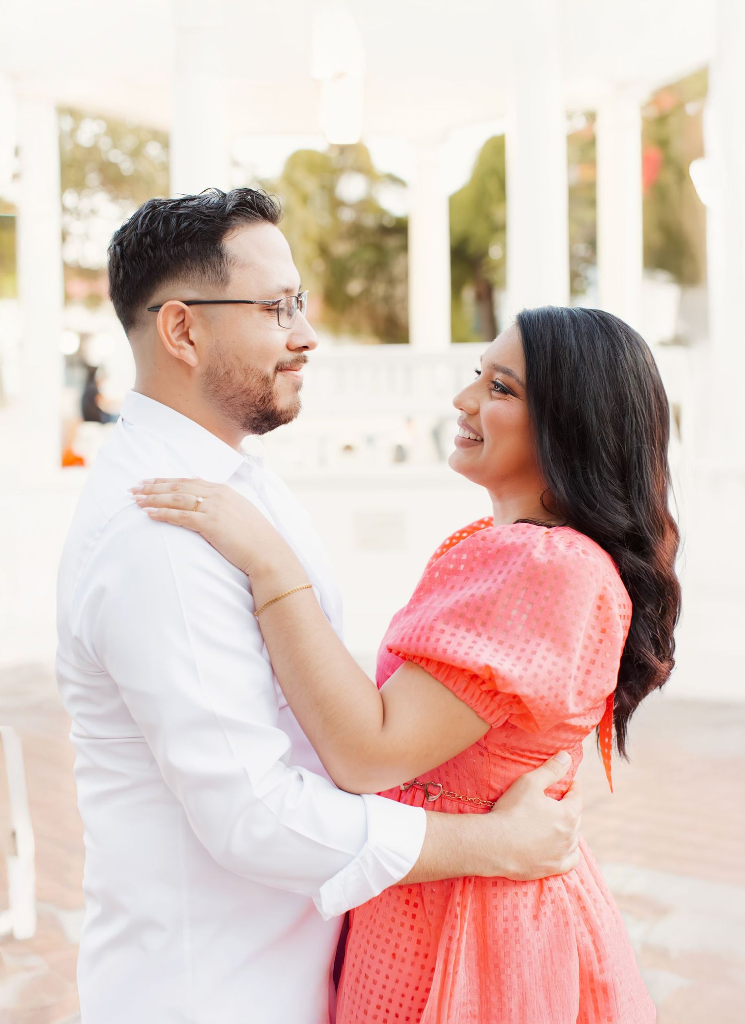 Happy couple in white and coral outfits embrace and smile during outdoor photoshoot.