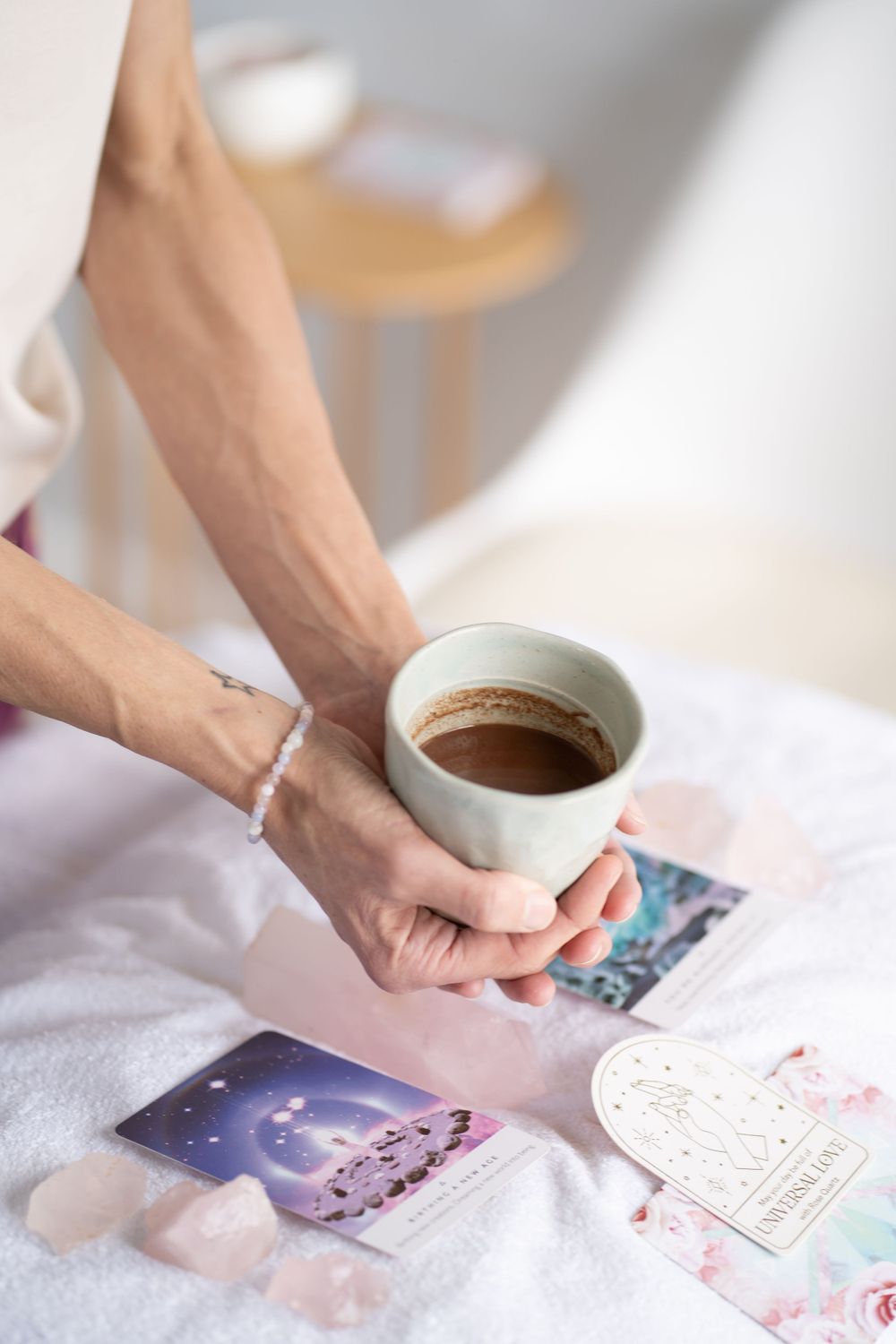 Hands holding a coffee cup over tarot cards and crystals on white fabric.