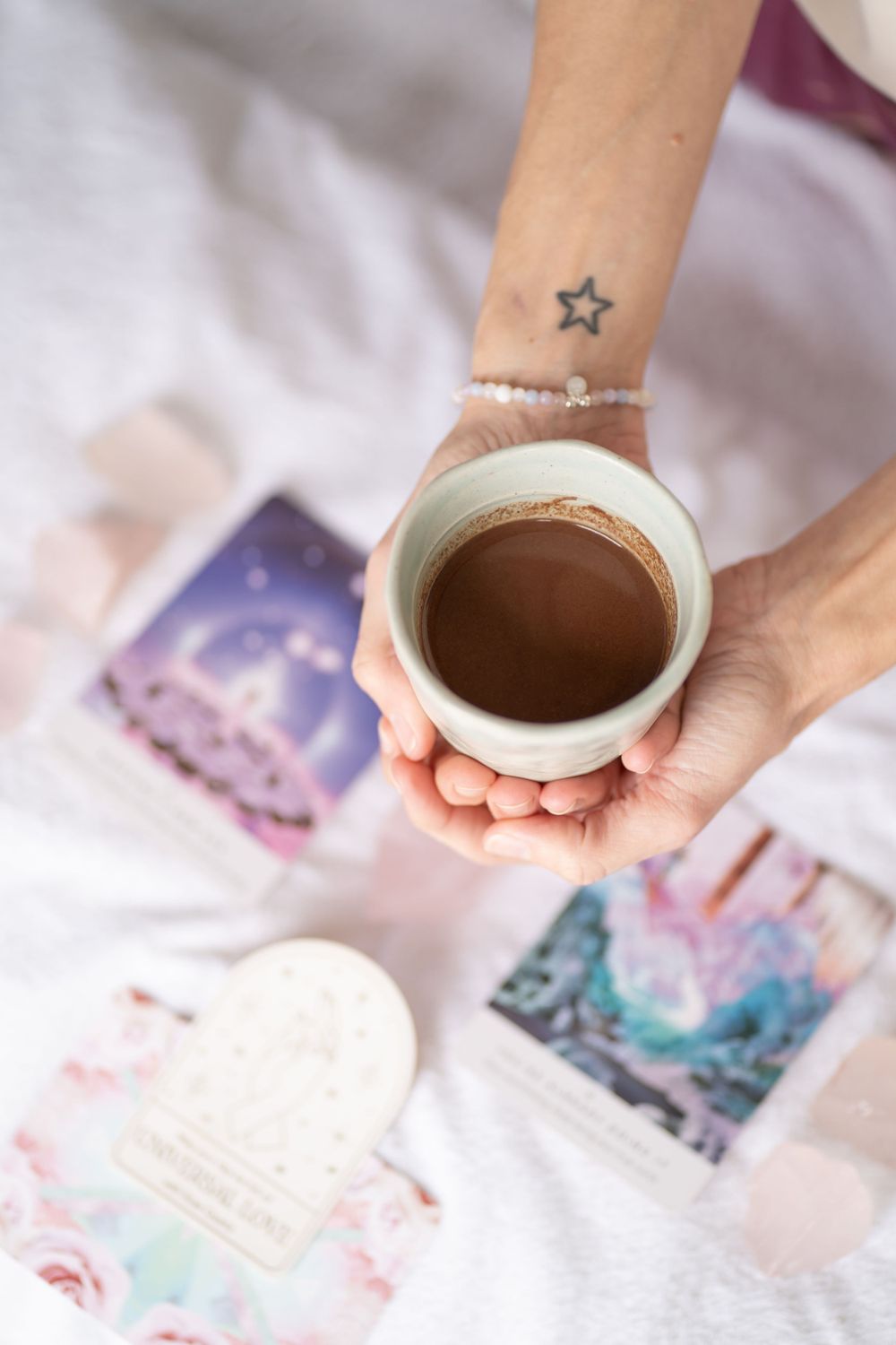 Close-up of hands holding coffee cup with small star tattoo and mystical cards visible.