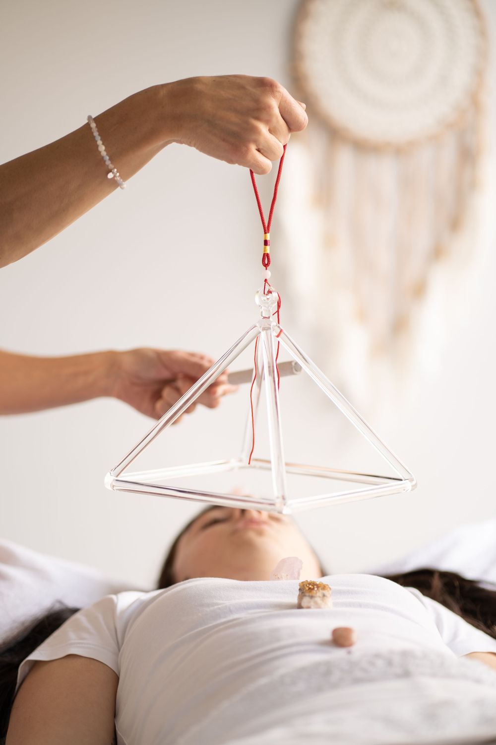 Hands holding a geometric crystal healing pyramid with dreamcatcher visible in background.