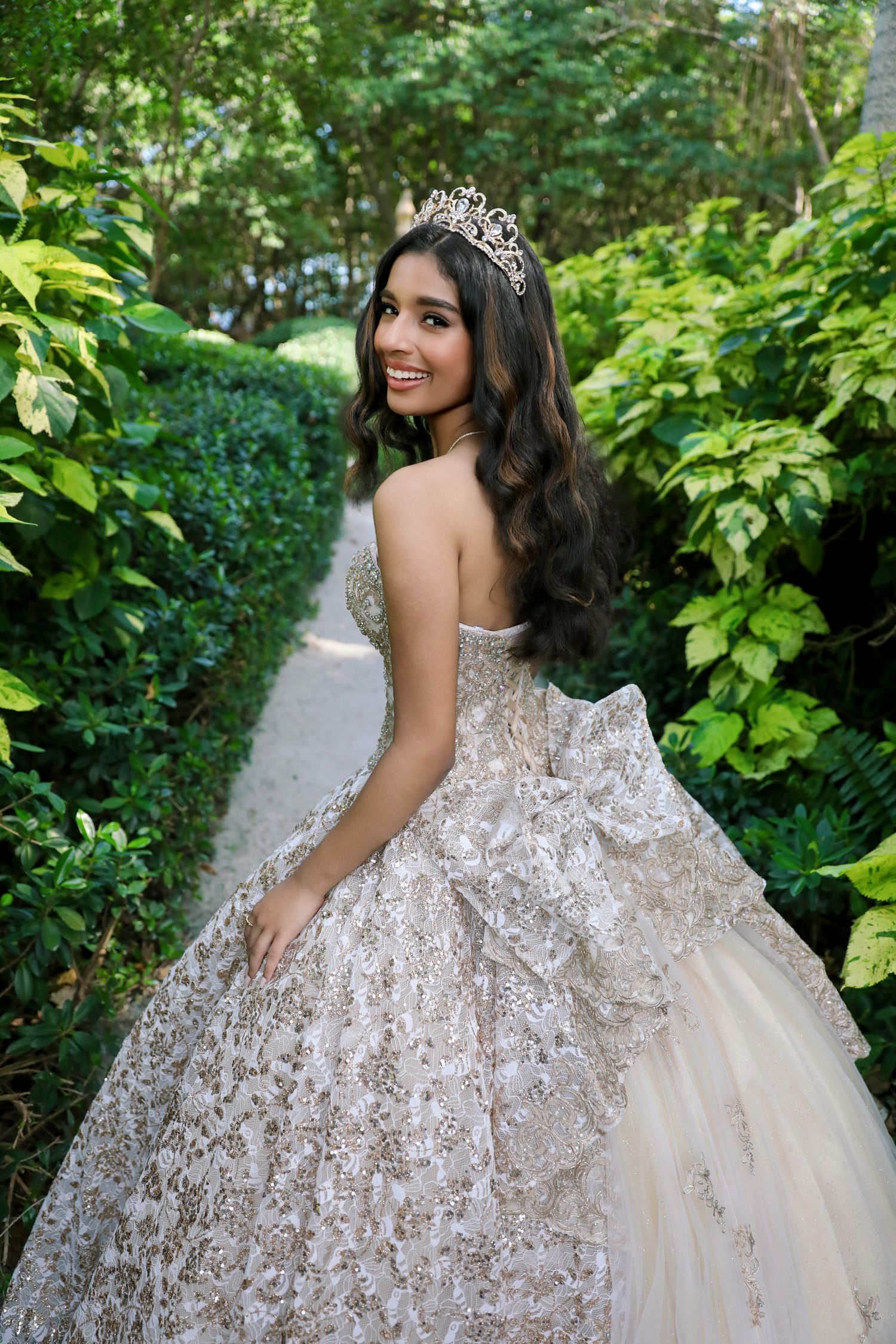 A princess-style portrait in an ornate ivory ball gown with lace details in a garden pathway.