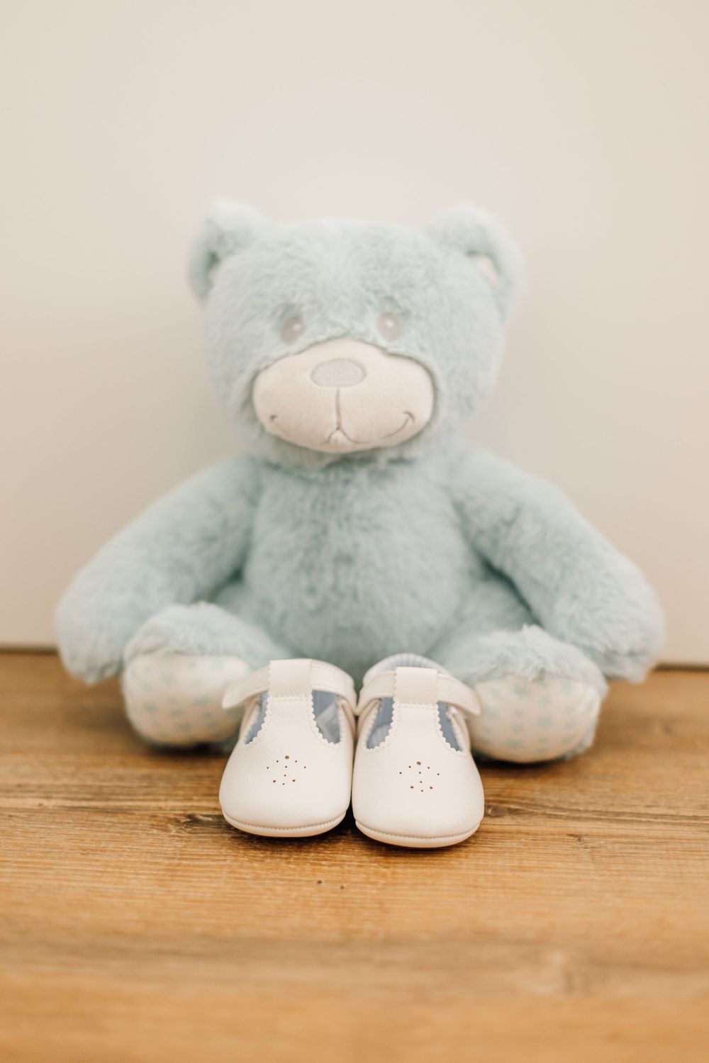 Light blue teddy bear with white baby shoes arranged on wooden floor.