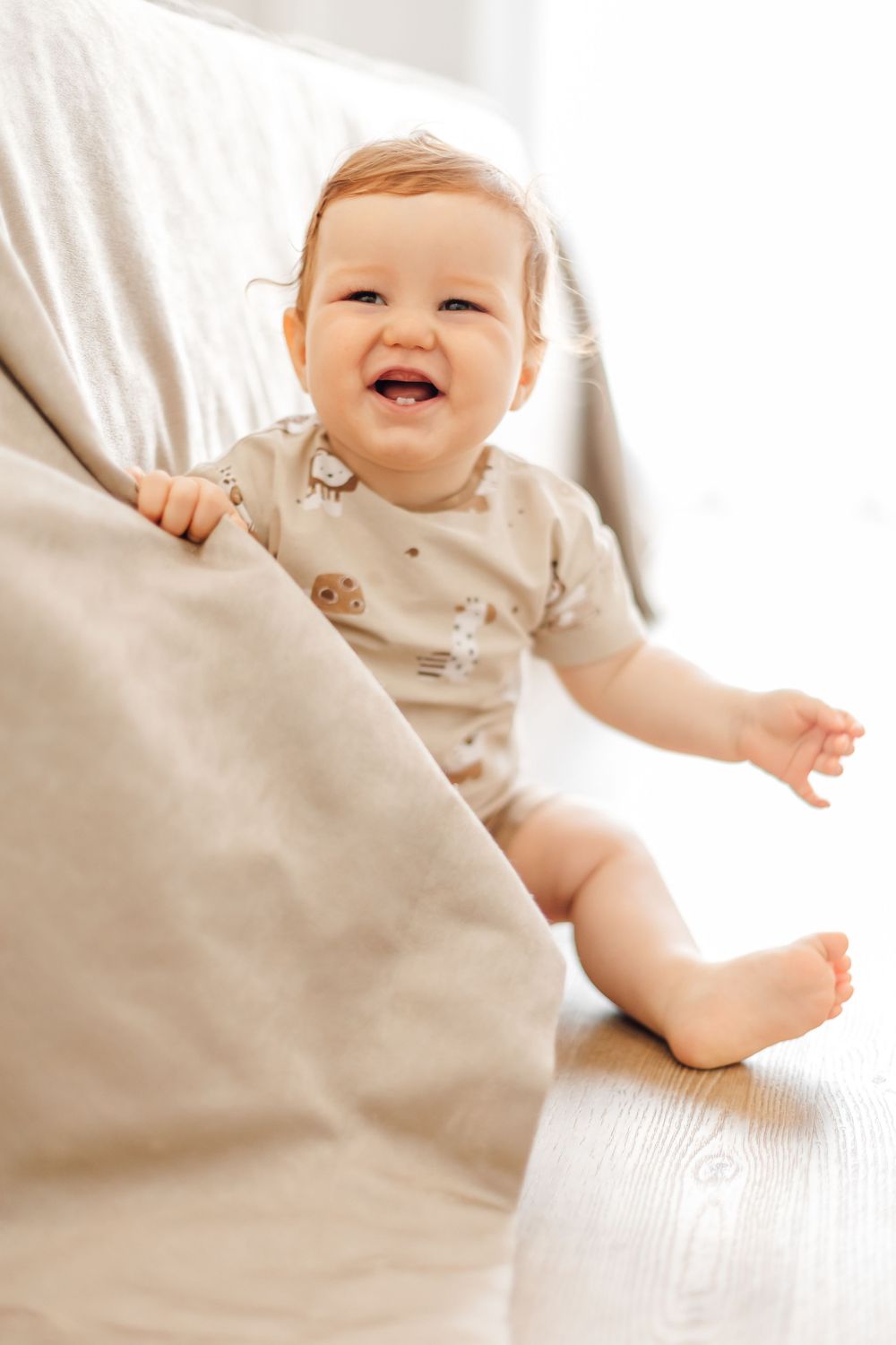 Happy baby in beige outfit sitting on white couch with soft cushions.
