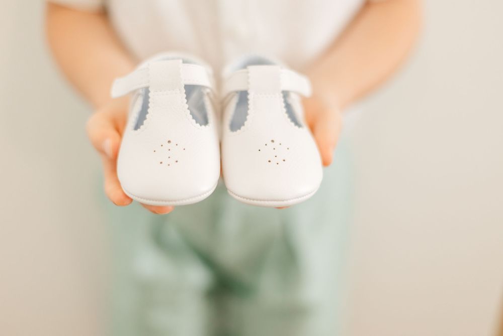 Small white baby shoes held in hands with a mint green background.