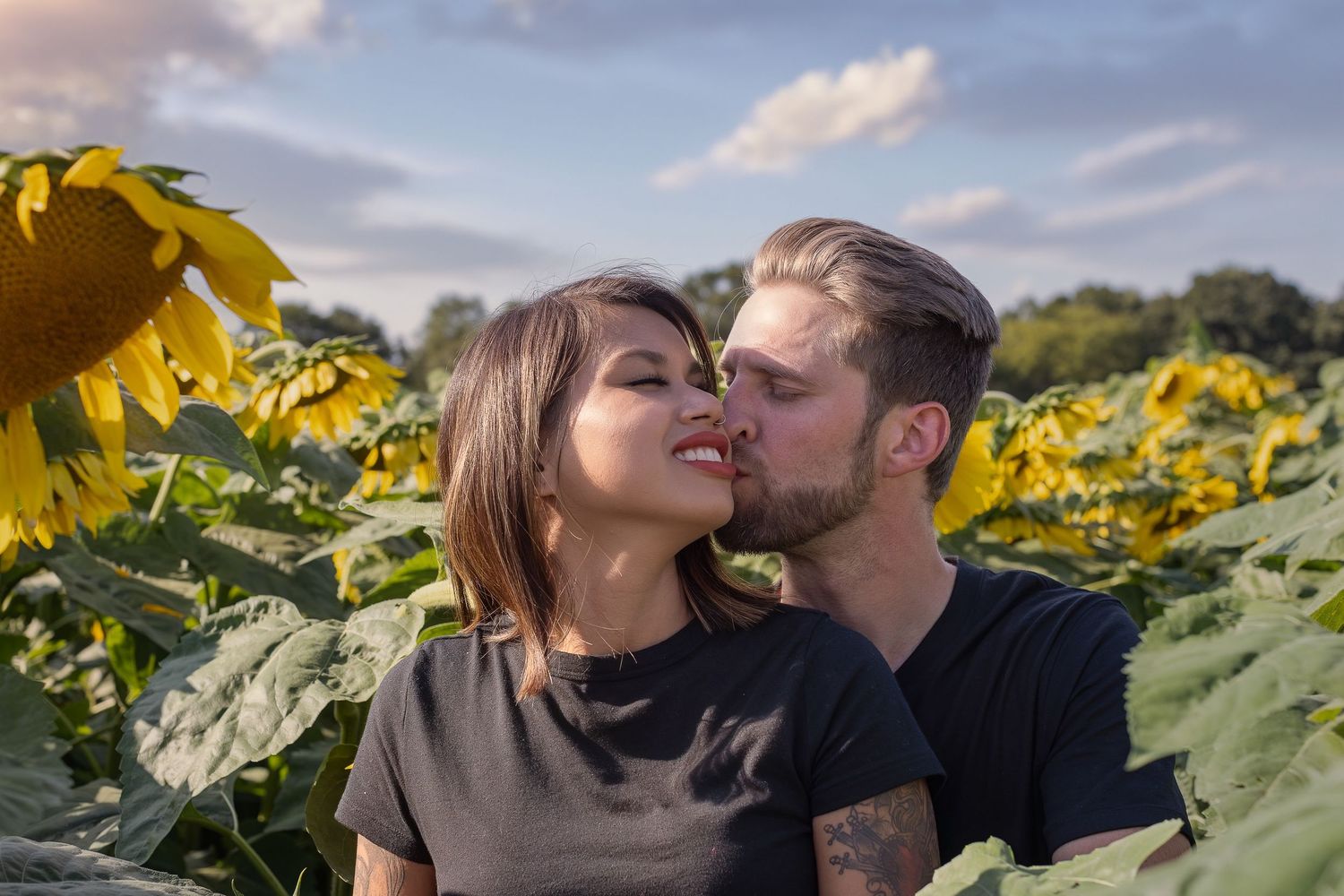 A couple embraces and kisses in front of a field of bright yellow sunflowers on a beautiful summer day.