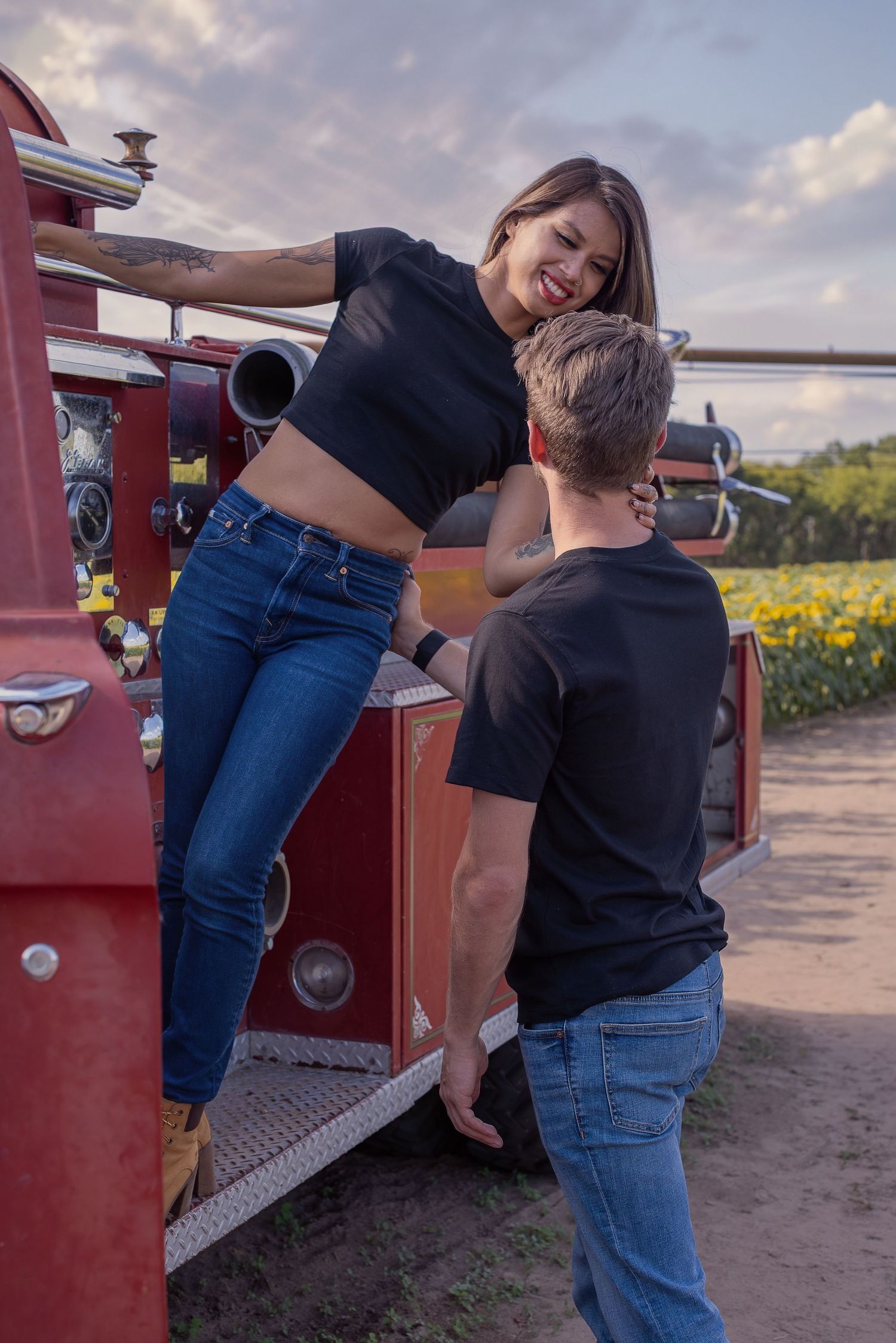 A playful couple laughs and interacts near an old red truck in a field of flowers.