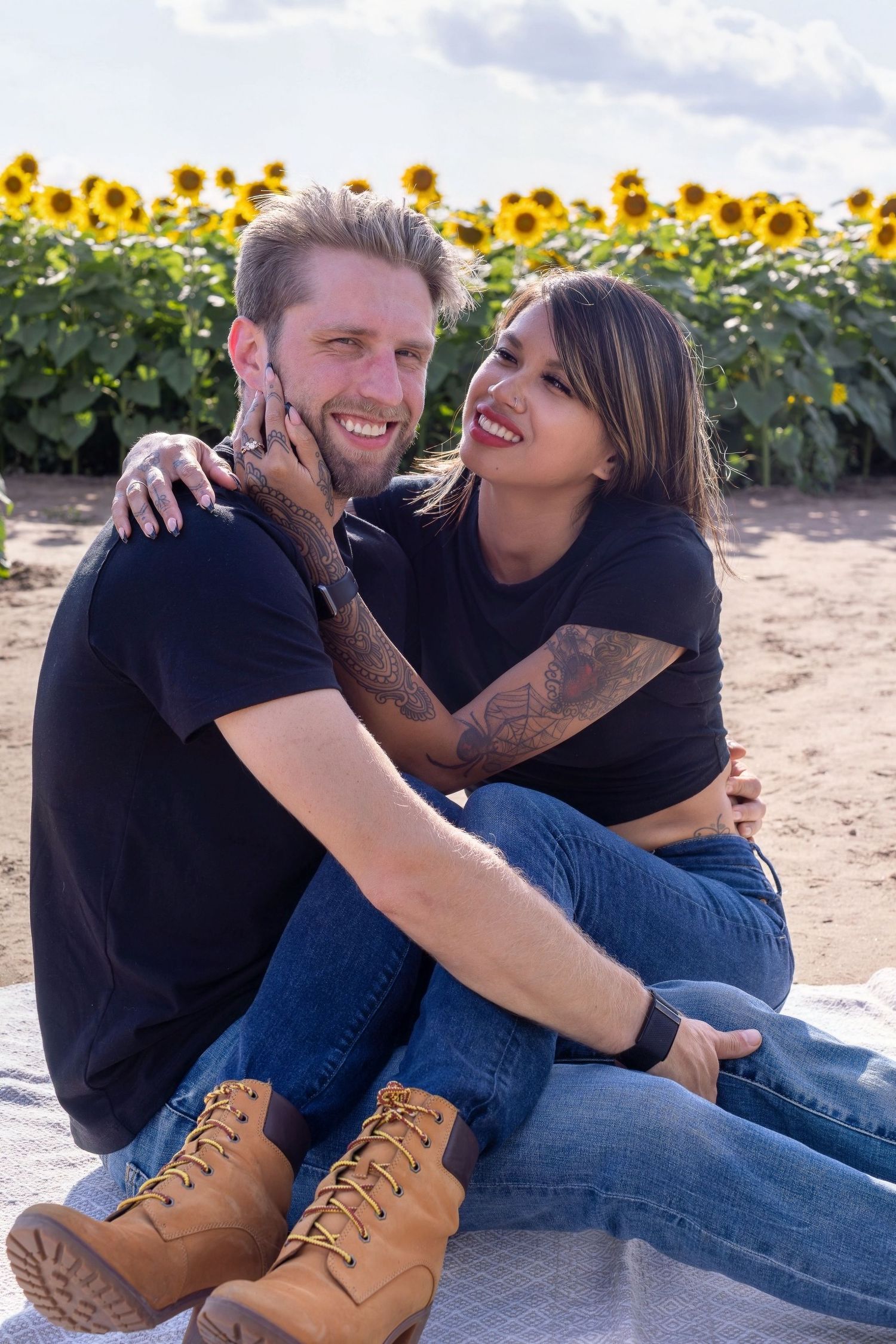 A casual couple laughing and embracing in a sunflower field wearing jeans and casual attire during a sunny outdoor photoshoot.