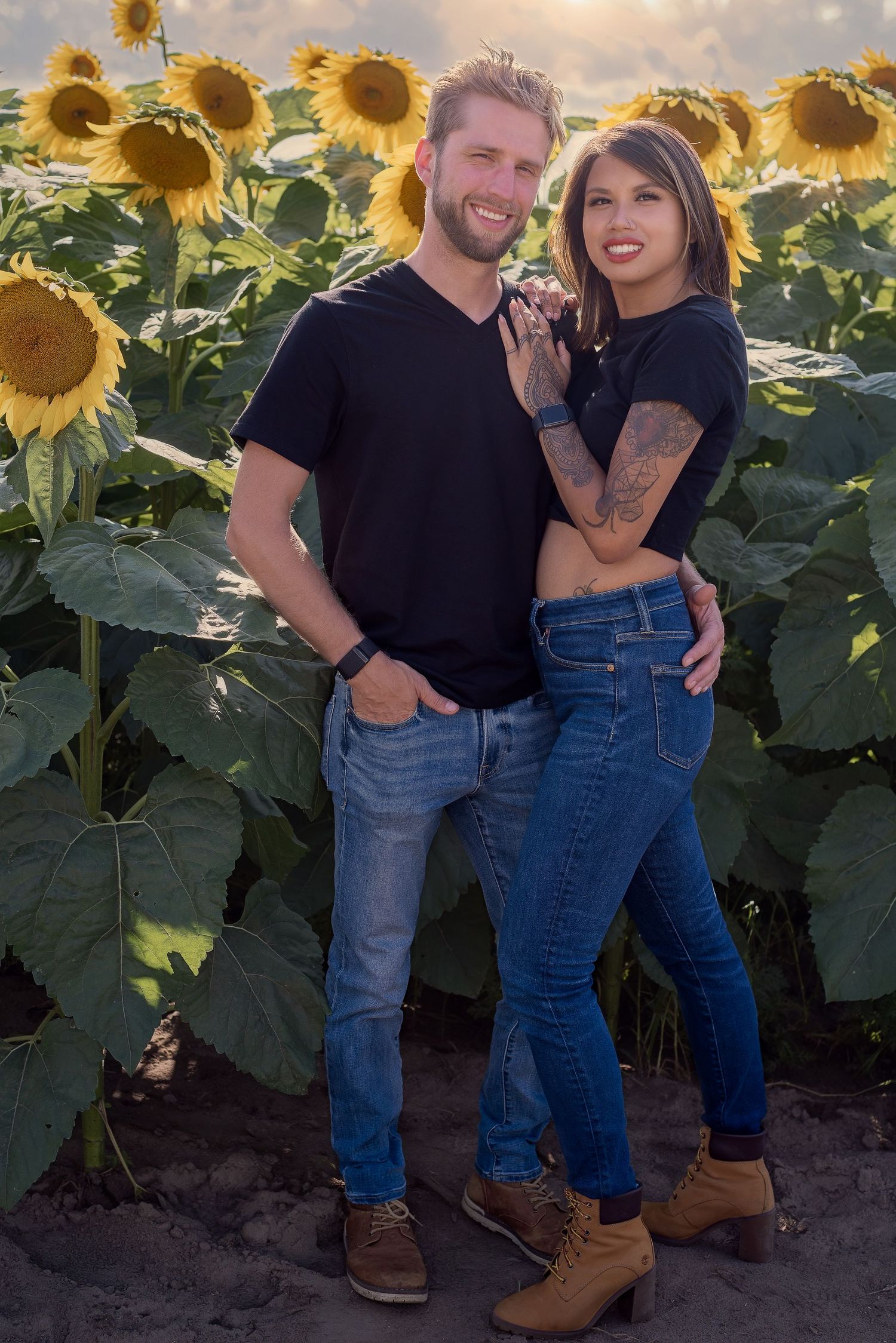A couple in matching black shirts and jeans embraces in a sunflower field during sunset.