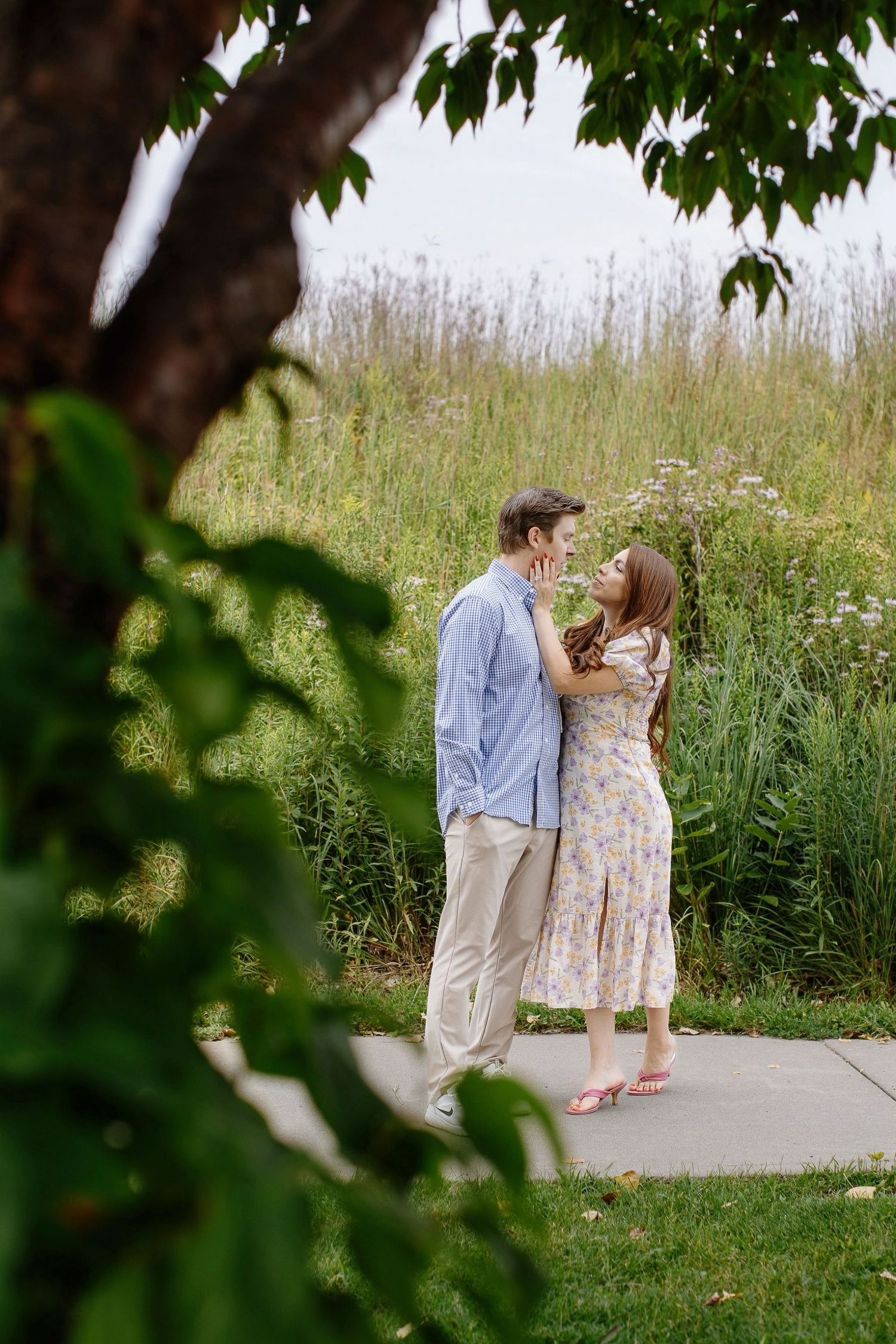A couple shares a romantic moment on a paved path surrounded by tall grasses and wildflowers on a summer day.