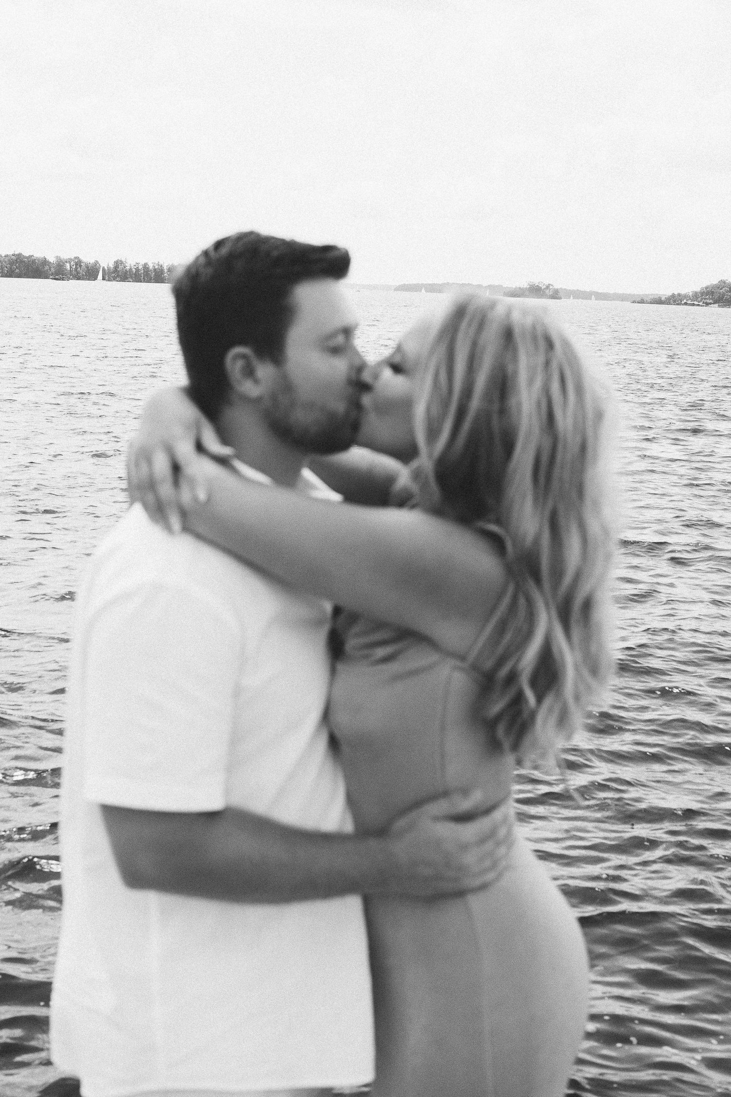 A romantic couple shares an intimate kiss while embracing by the water in a black and white photograph.