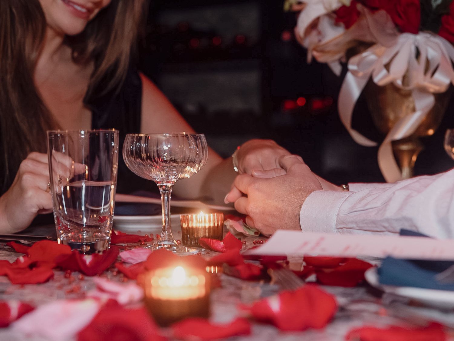 Romantic dinner table setting with glowing candles and scattered red rose petals on white tablecloth.