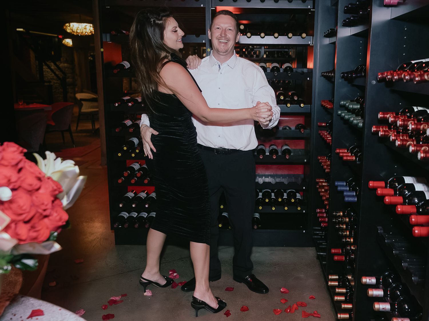 A couple dances together in a dimly lit venue with wine racks visible in the background.