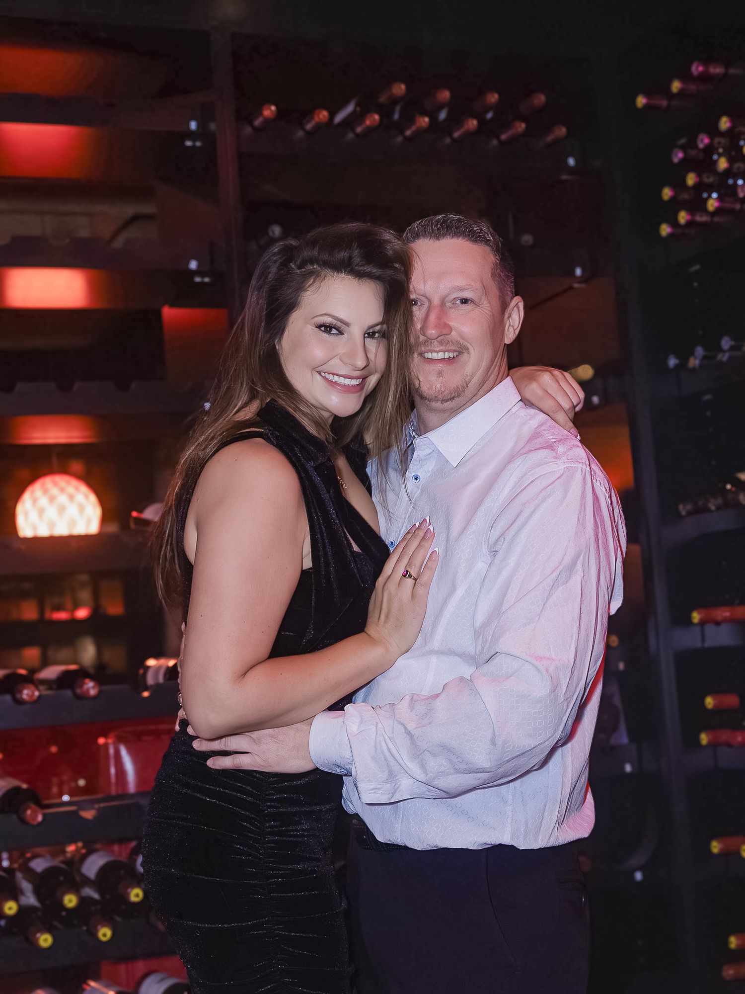 A couple embraces on a dance floor at a dimly lit venue with festive lights in the background.