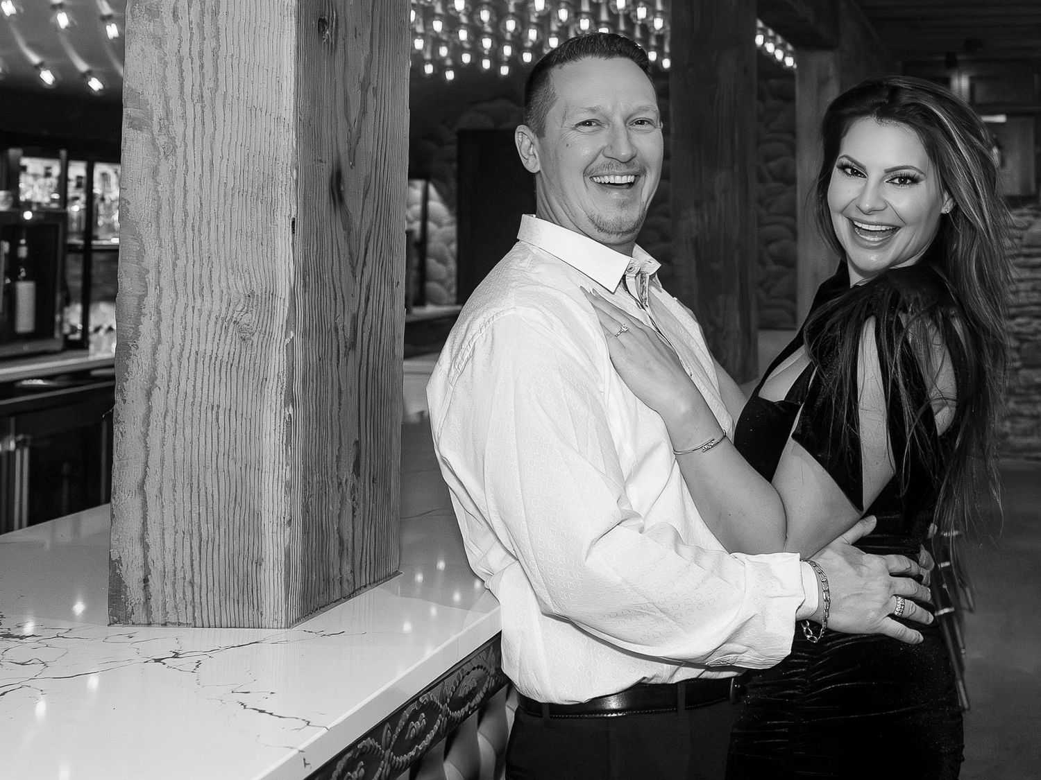 A couple shares a playful dance moment in front of wooden beams and string lights.