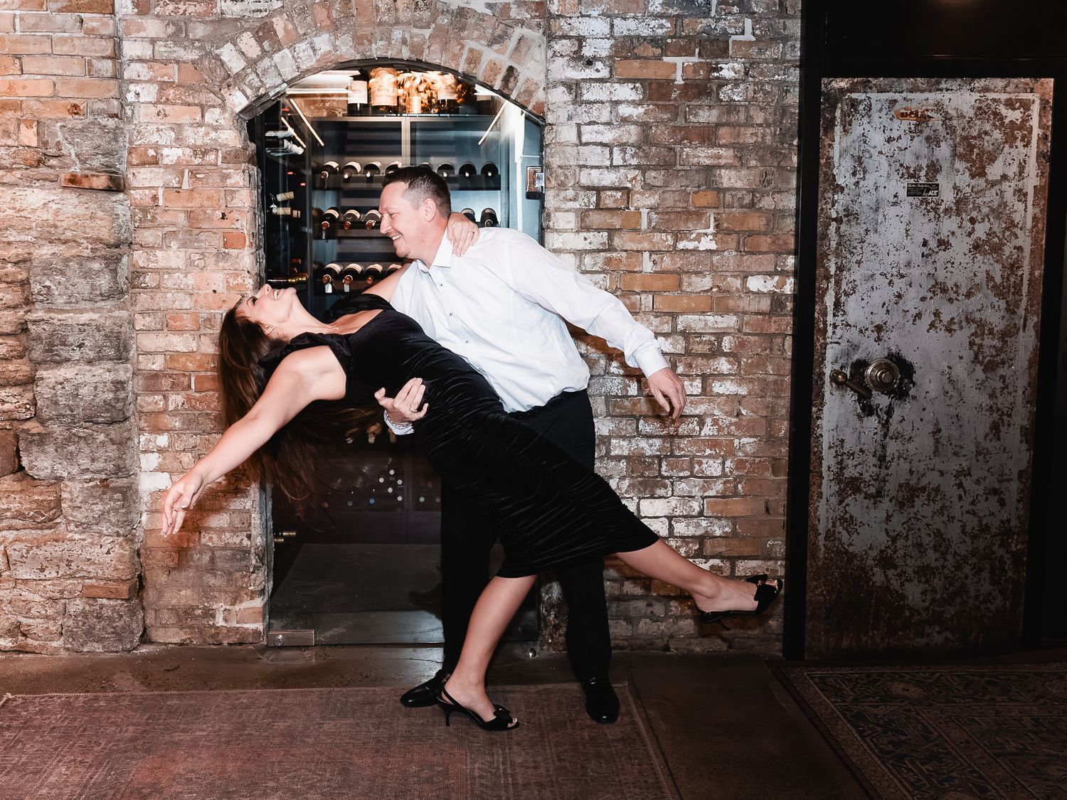 A couple performs a dramatic dance dip pose against an exposed brick wall background.