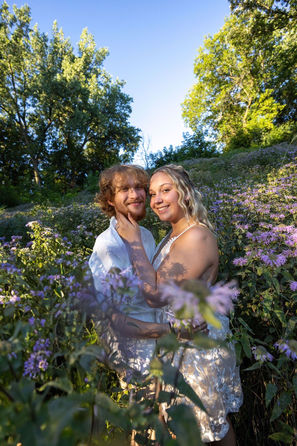 Couple embracing among purple wildflowers in sunny garden setting.