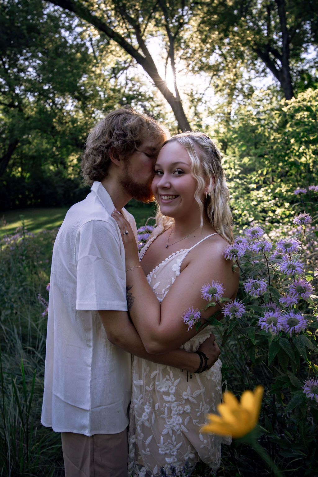 Romantic couple embraces in a garden with purple wildflowers during golden hour sunset.