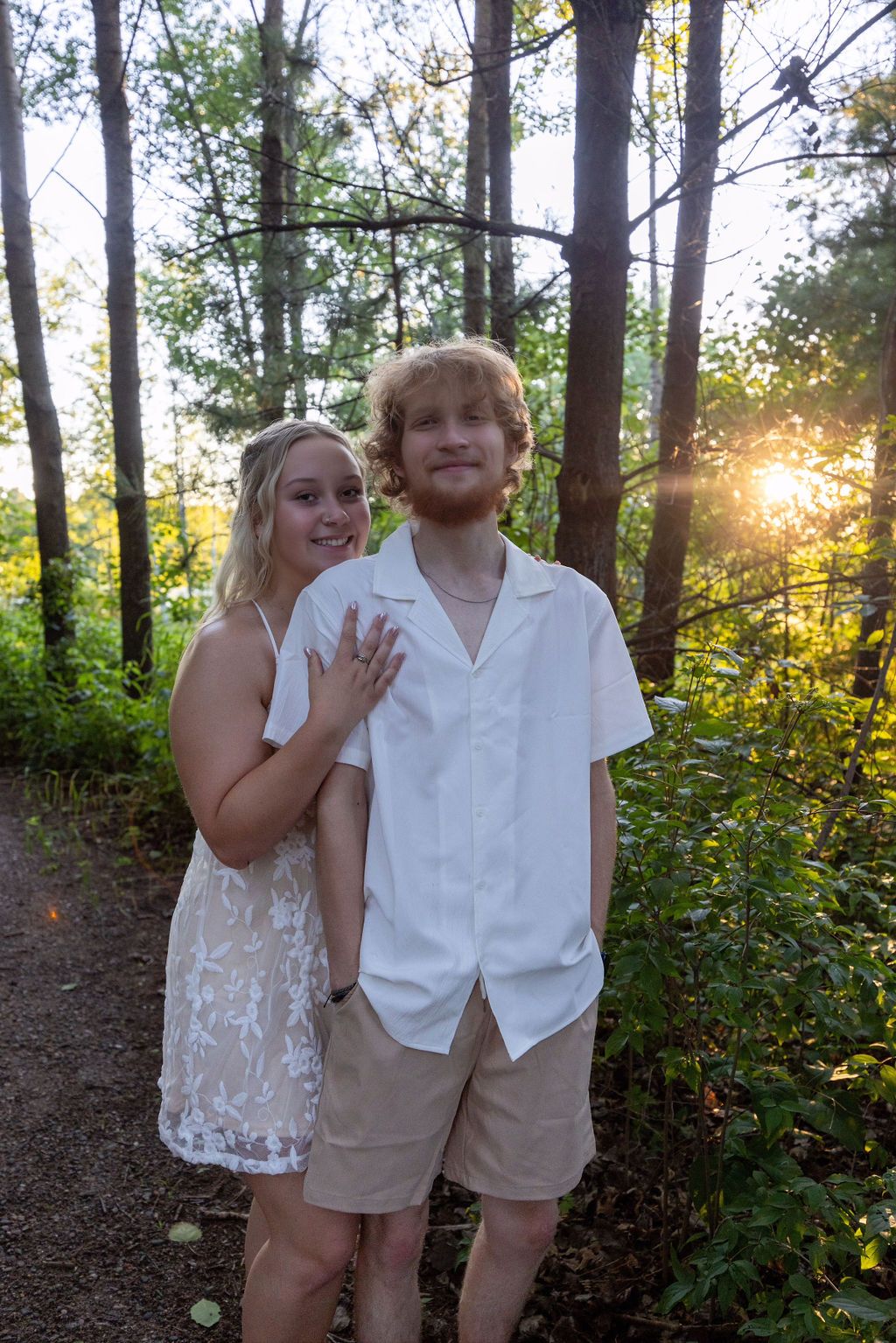 A couple shows off an engagement ring while standing on a forest path at sunset.