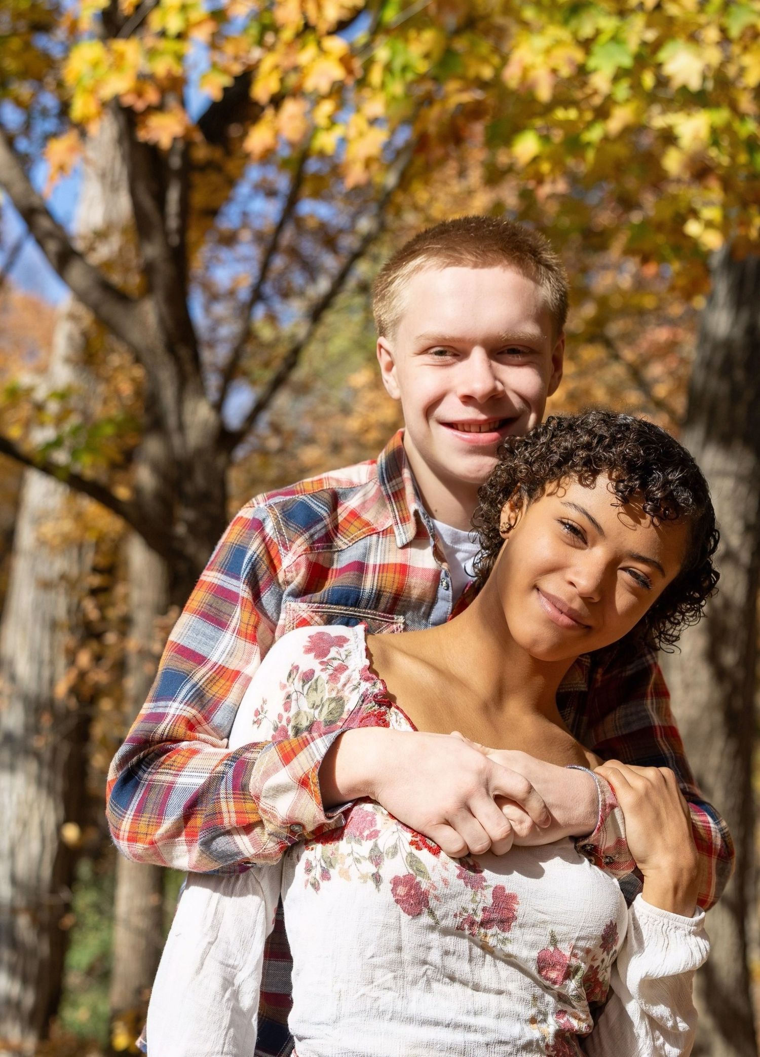 A romantic couple embraces while standing in front of autumn trees with golden leaves.