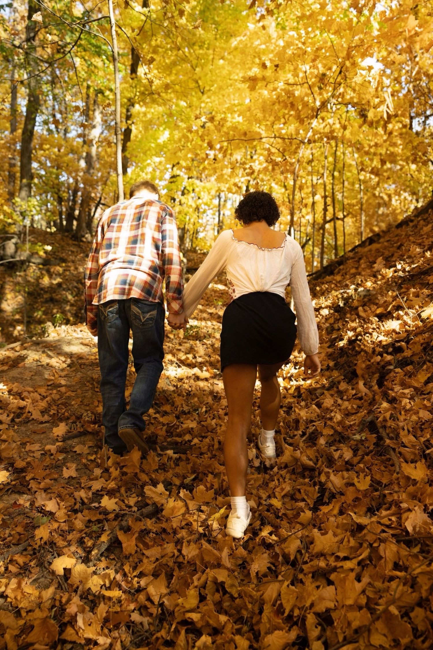 A couple walks up a leaf-covered hill surrounded by golden autumn trees.