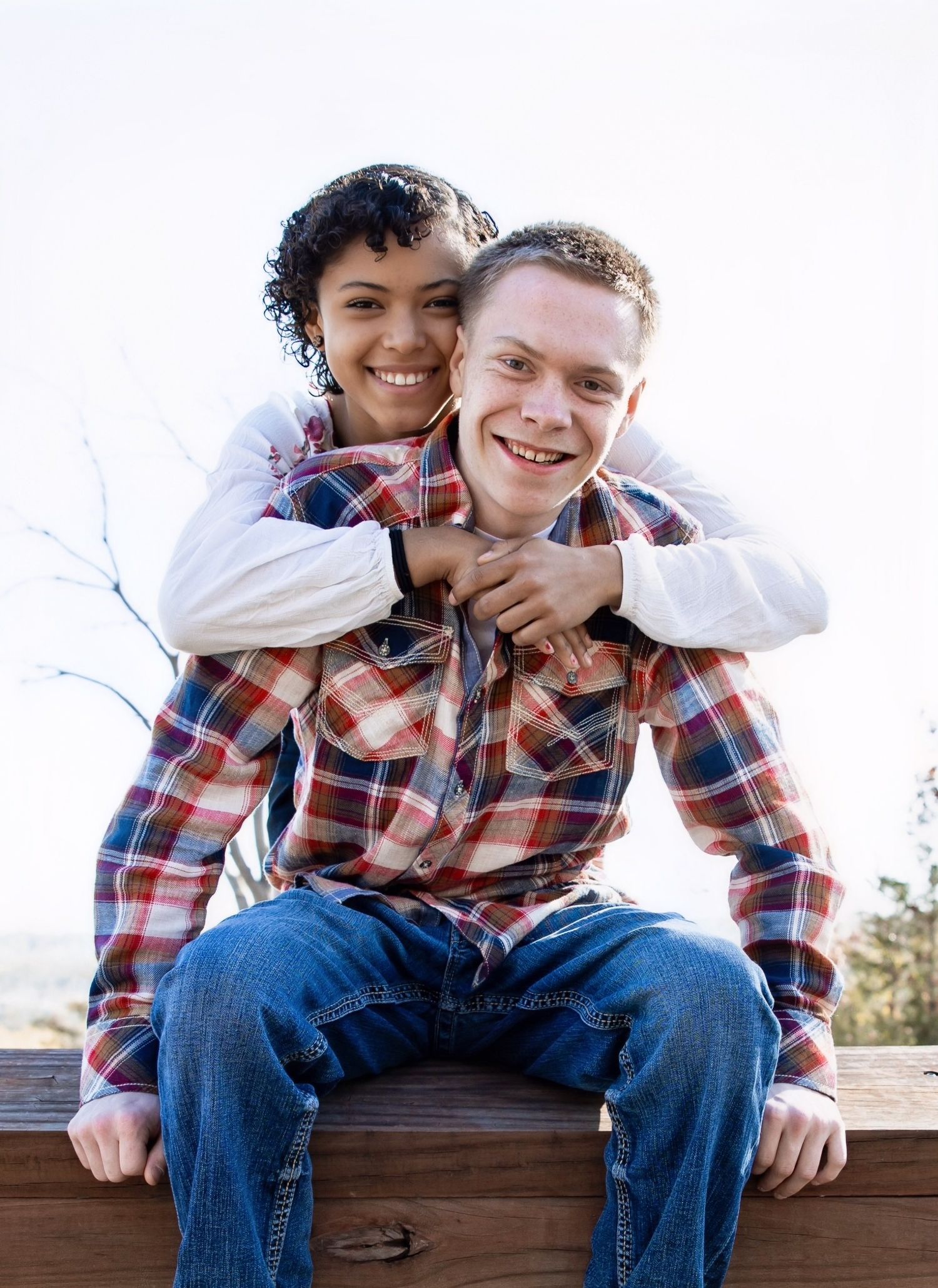 A happy couple shares a playful piggyback moment while sitting on a wooden railing outdoors.