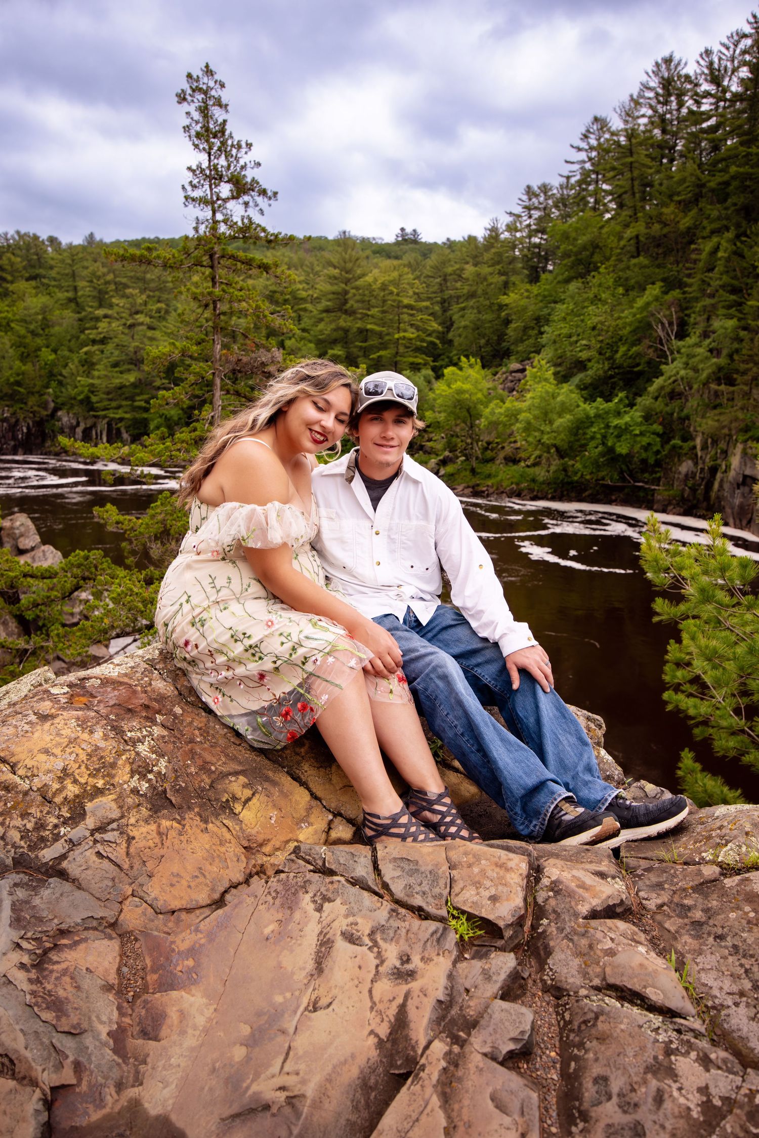 A couple sits together on a rocky outcrop overlooking a scenic river surrounded by lush forest and dramatic cliffs.